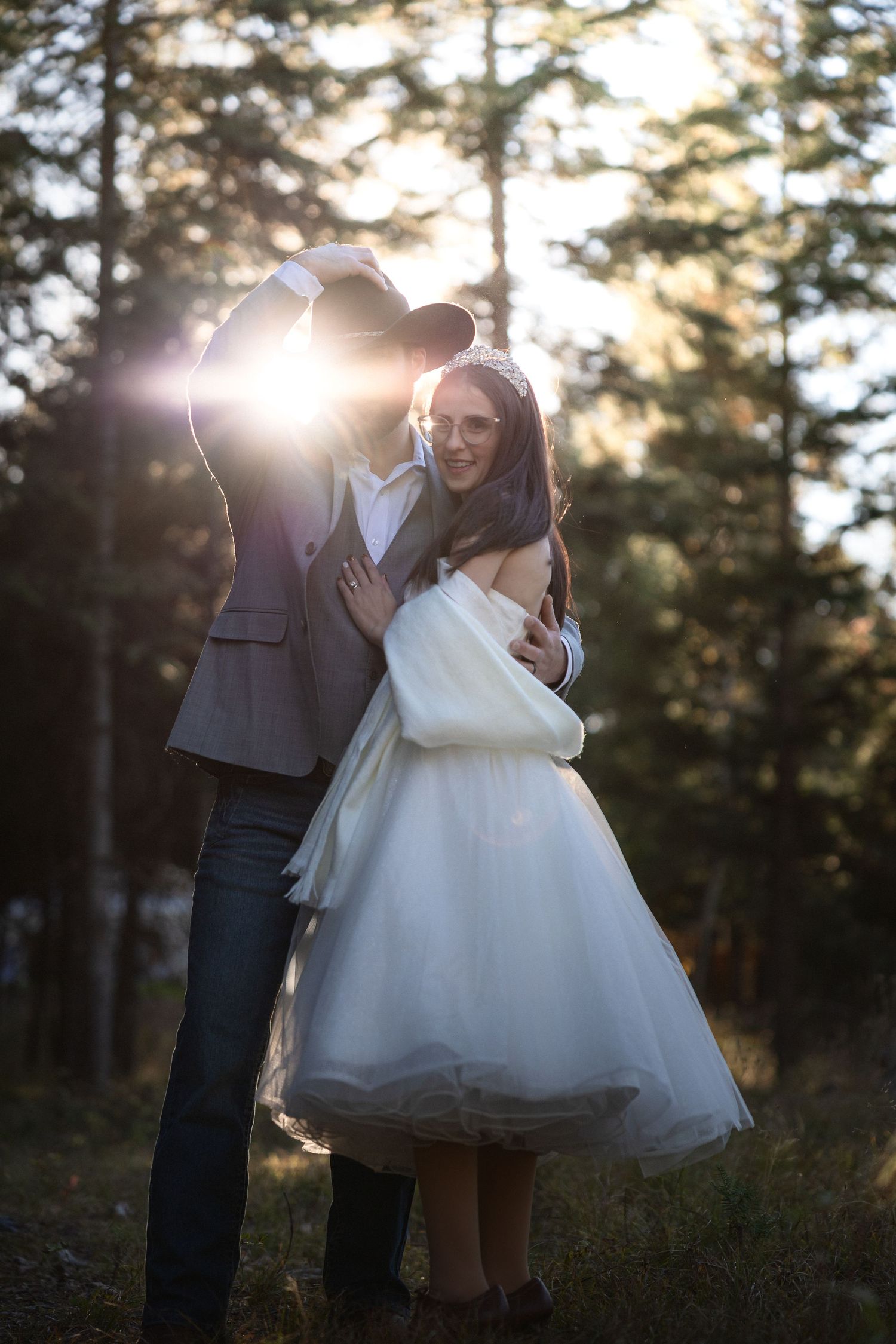 A tender wedding moment backlit by sunlight filtering through pine trees in a forest.