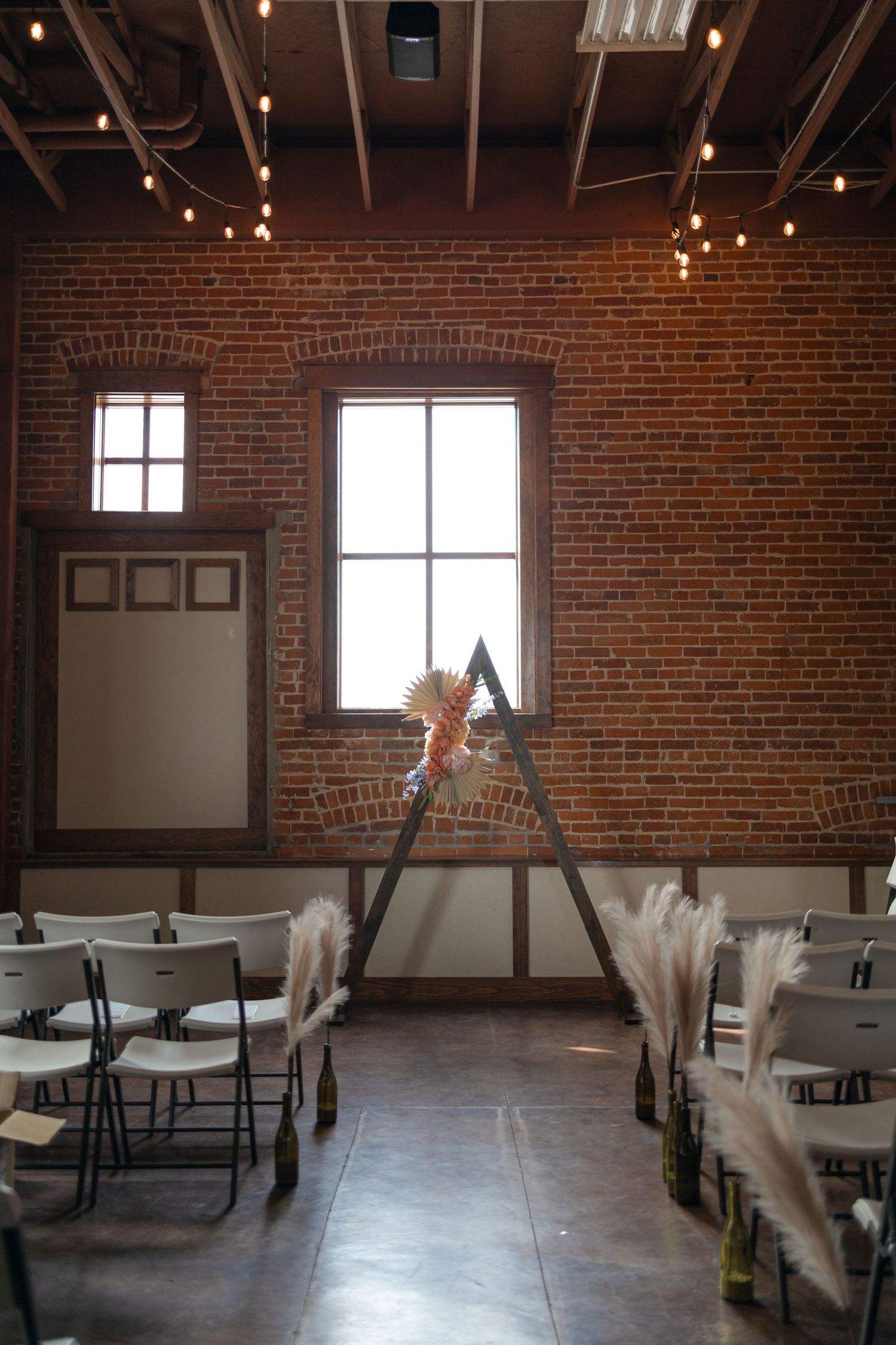 Wedding ceremony setup with triangle arch and string lights in industrial brick venue.