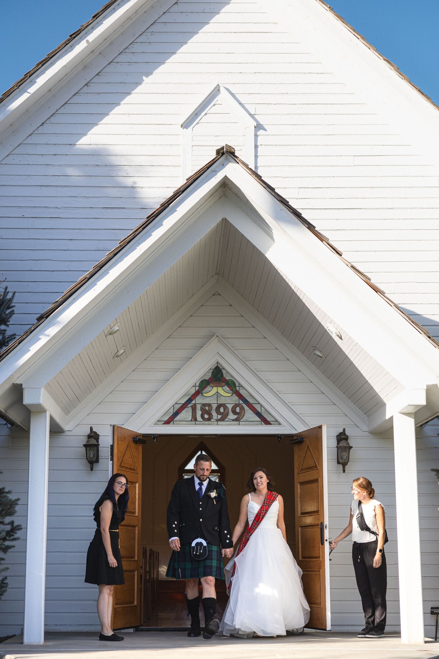 Wedding party standing at entrance of historic white chapel with 1899 marking above door.