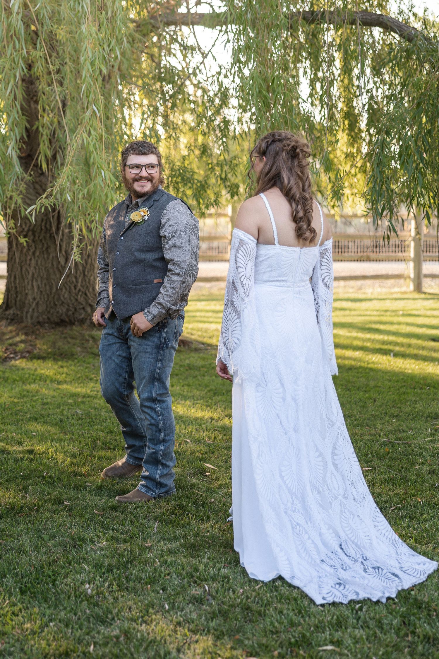A couple poses together in a grassy outdoor setting with willow trees, the bride wearing a flowing white lace dress.