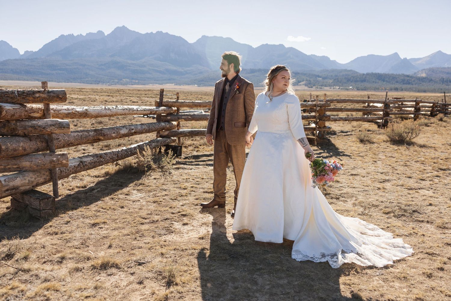 A wedding couple embraces in front of a rustic wooden fence with mountain vistas in the distance.