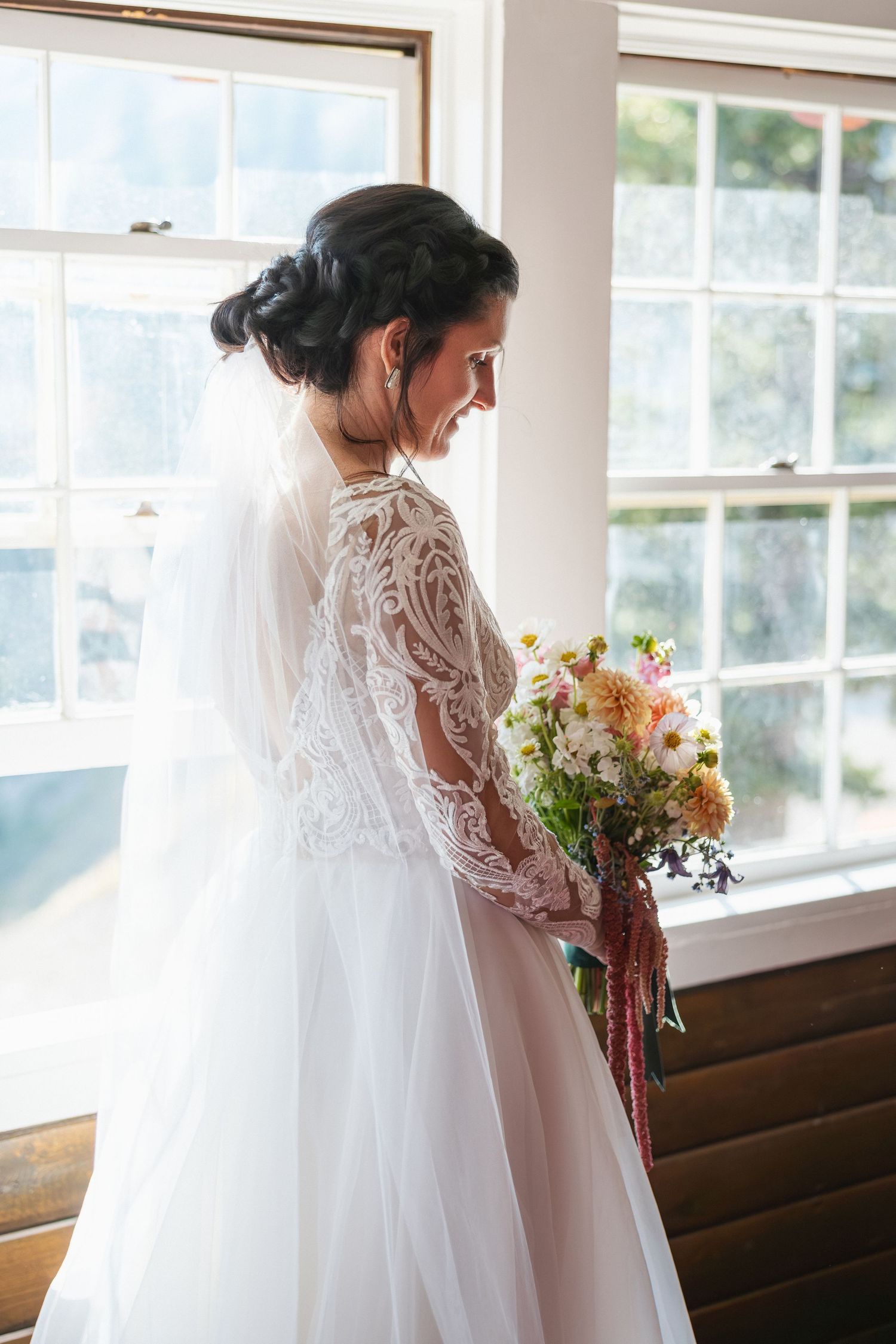 Bride holding autumn bouquet by sunlit windows in rustic venue.
