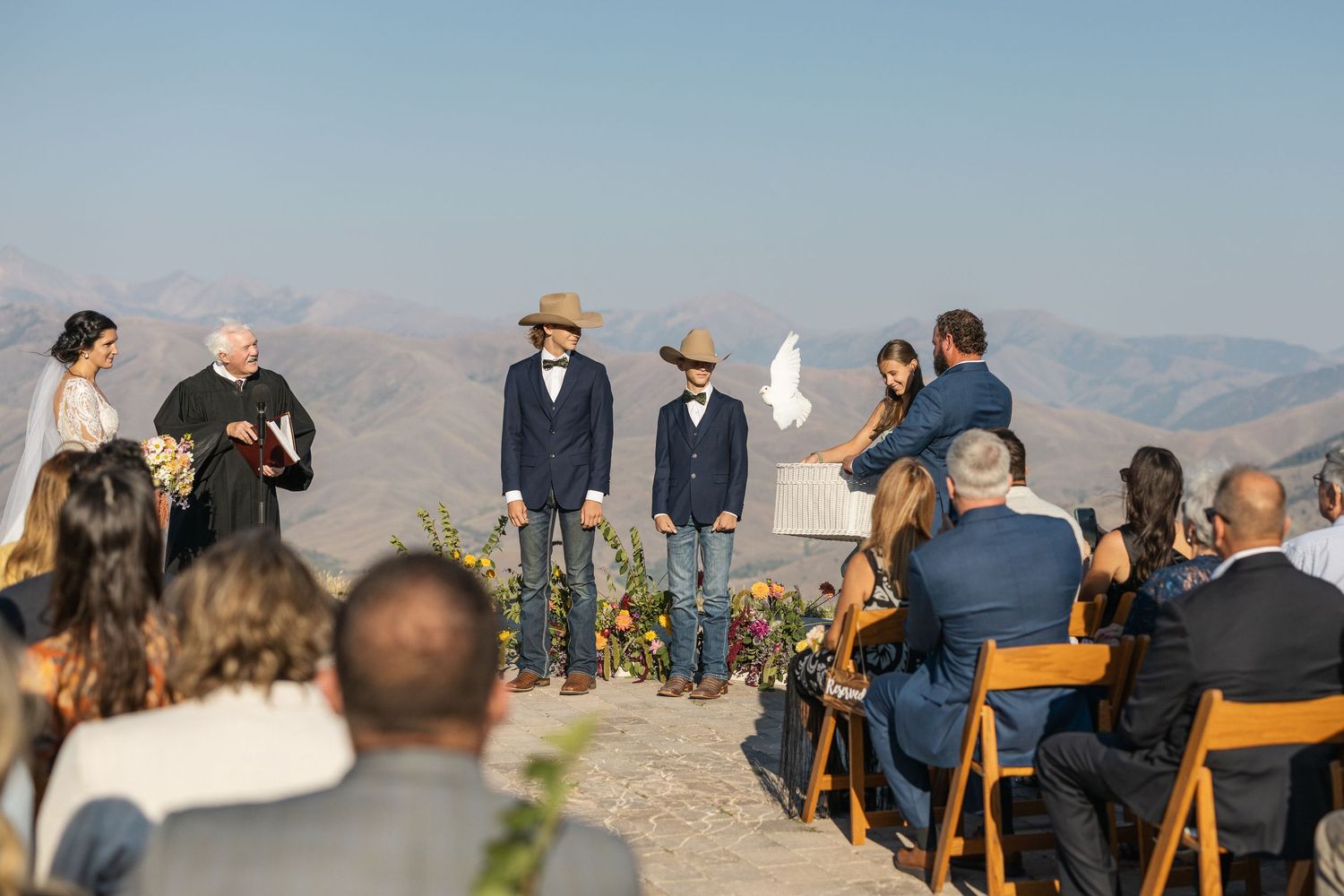 An intimate mountain wedding ceremony with guests seated on wooden chairs against a backdrop of rolling hills.