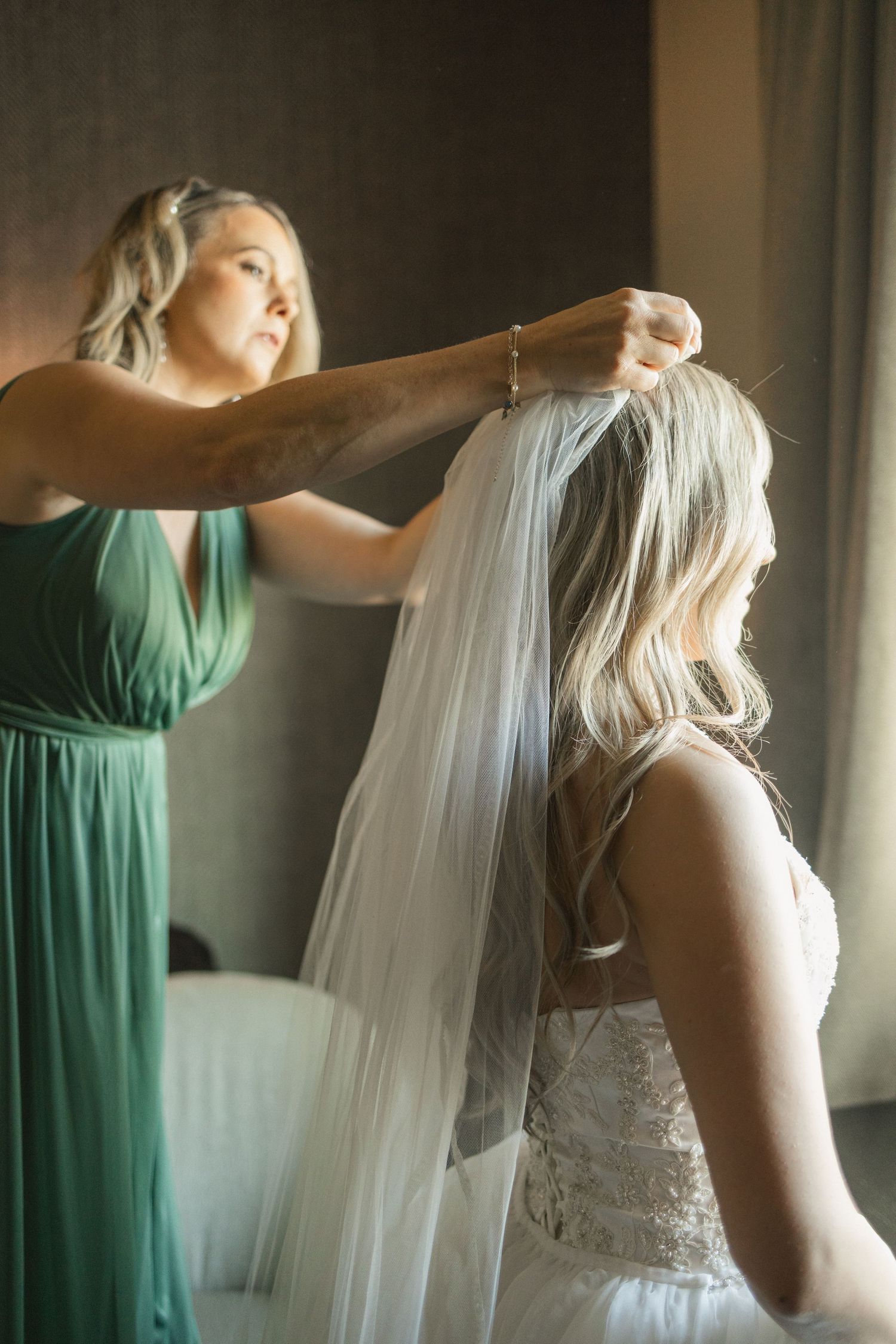 Bridesmaid in green dress helping attach bridal veil to bride's blonde hair.