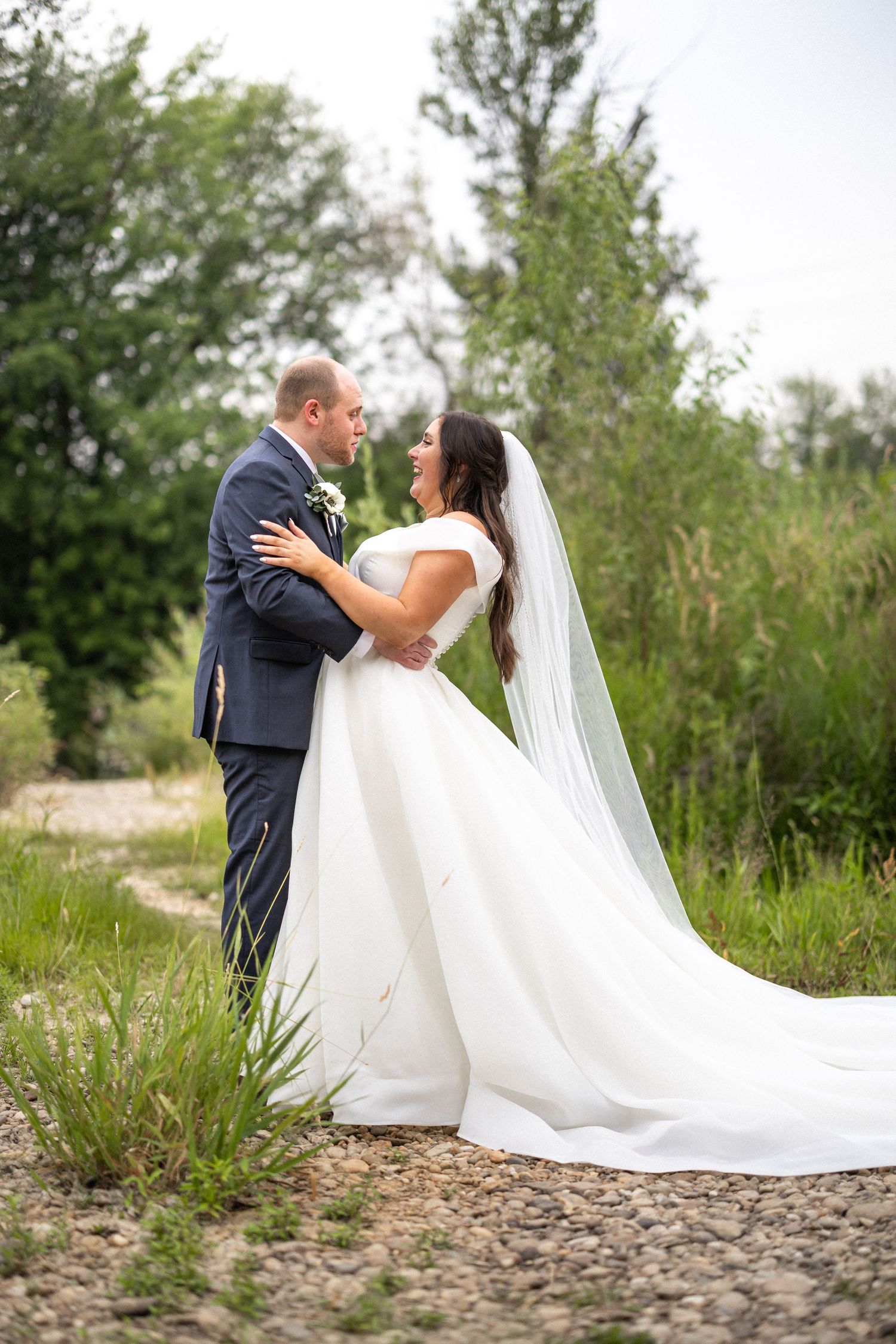 Romantic wedding couple embrace on nature path with bride's dress trailing behind.