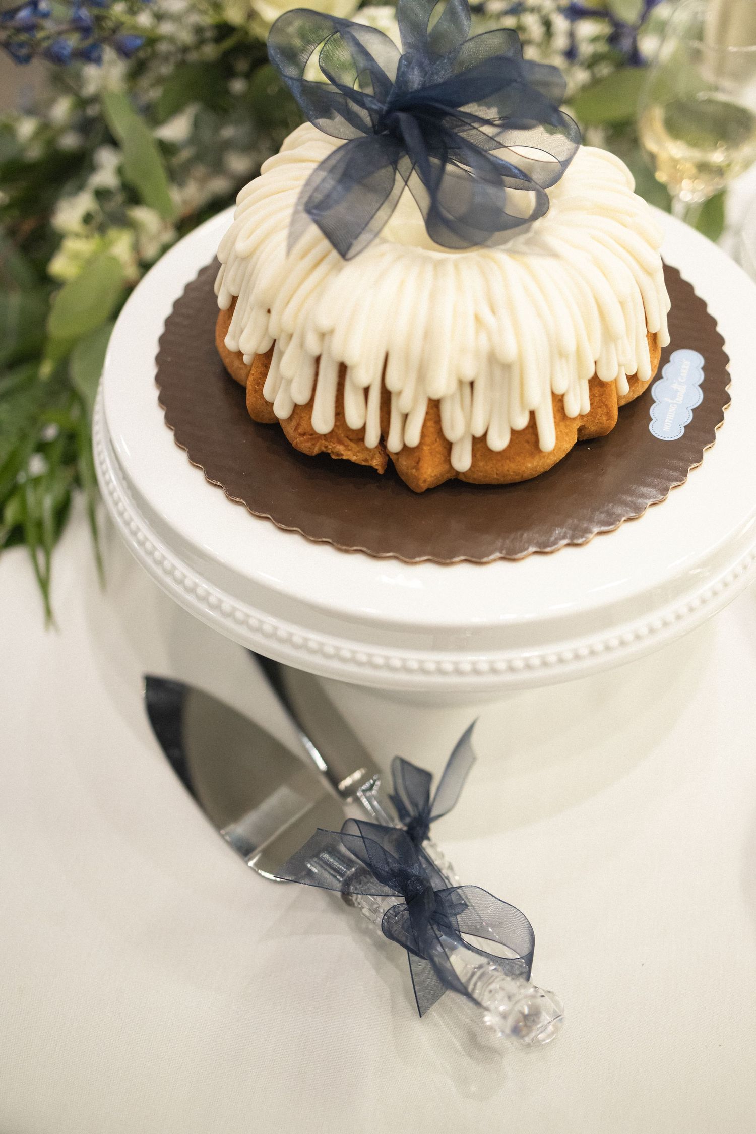 Elegant bundt cake with white drizzle and navy blue bow on a white pedestal cake stand.