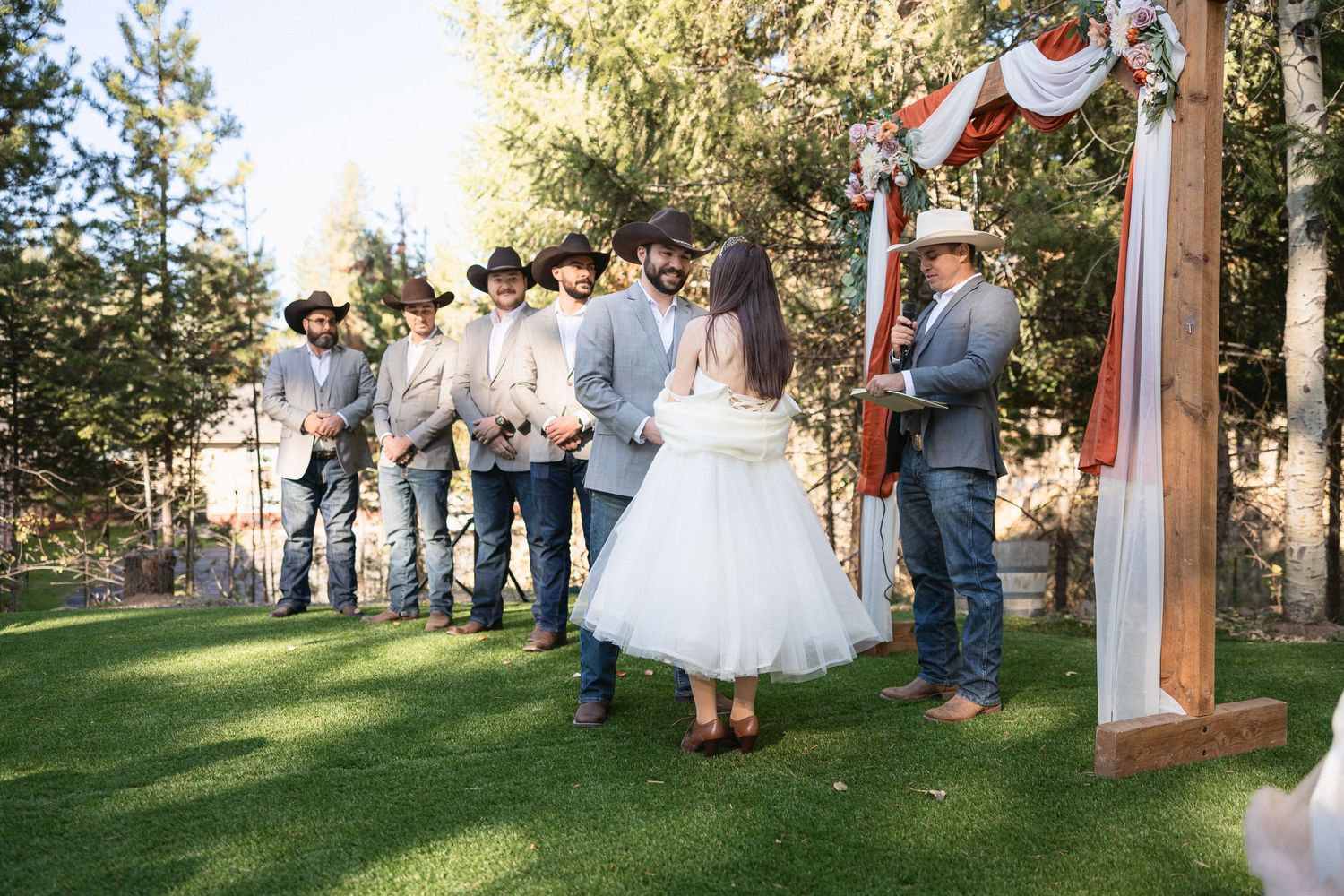 Wedding ceremony with groomsmen in cowboy attire standing near decorated wooden arch.