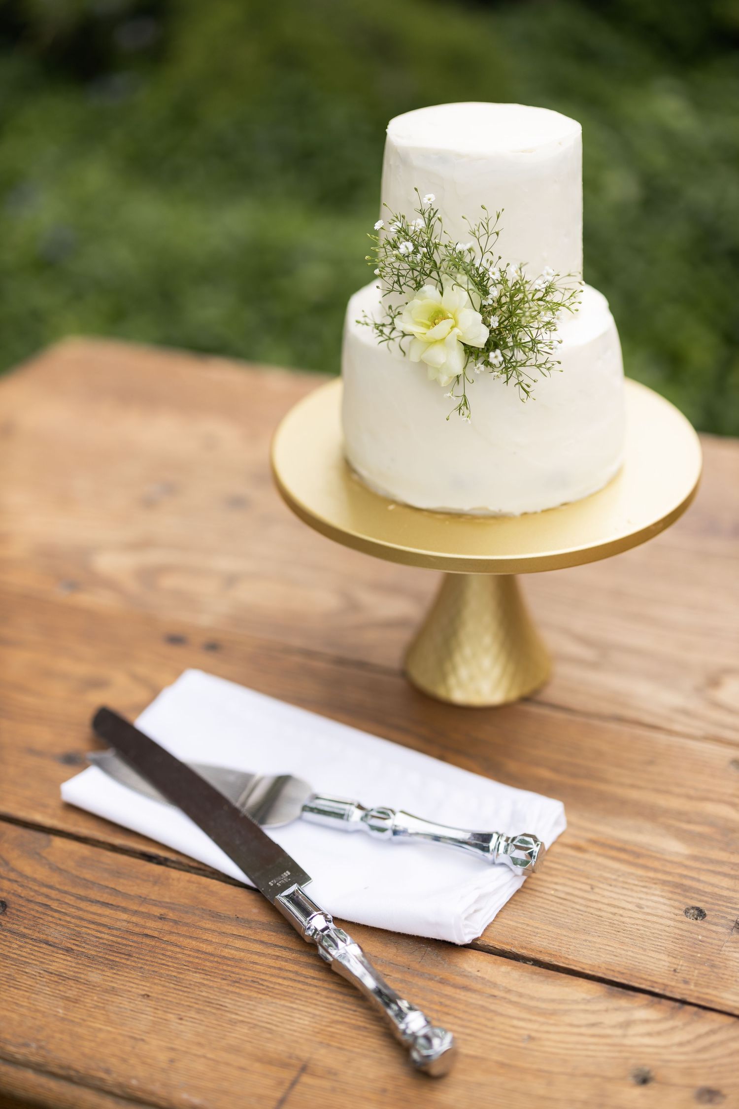 A simple two-tier white wedding cake with floral decoration sits on a gold stand atop a wooden table.