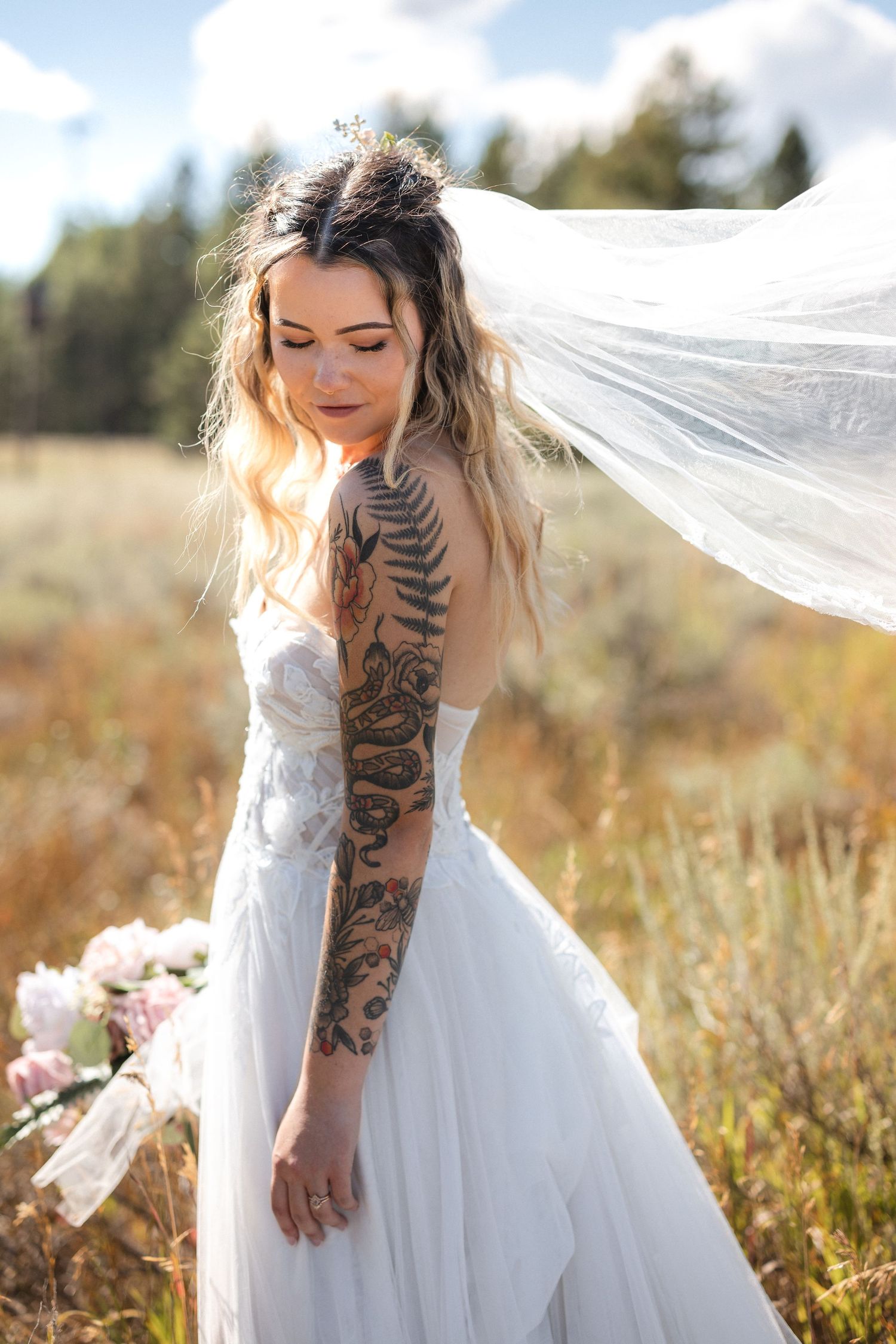 A bride in a strapless white gown with intricate arm tattoo poses in a sun-drenched field.