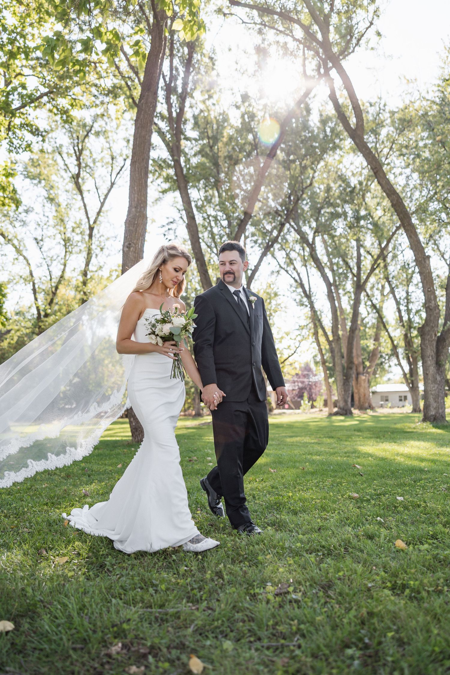 Couple in wedding attire walks through a sunlit park with flowing veil and trees in the background.