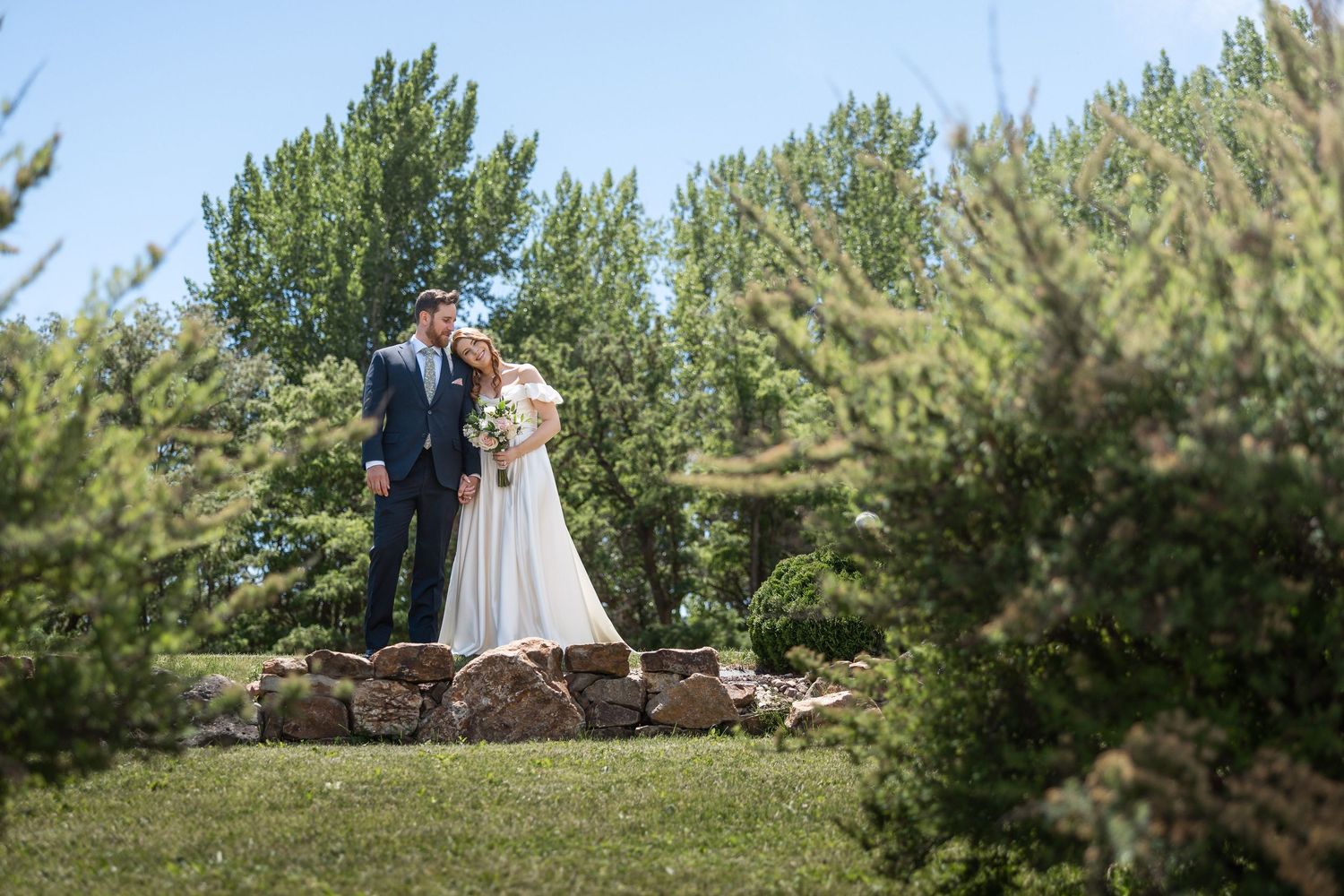 A couple stands on a stone wall surrounded by pine trees in a scenic outdoor setting.