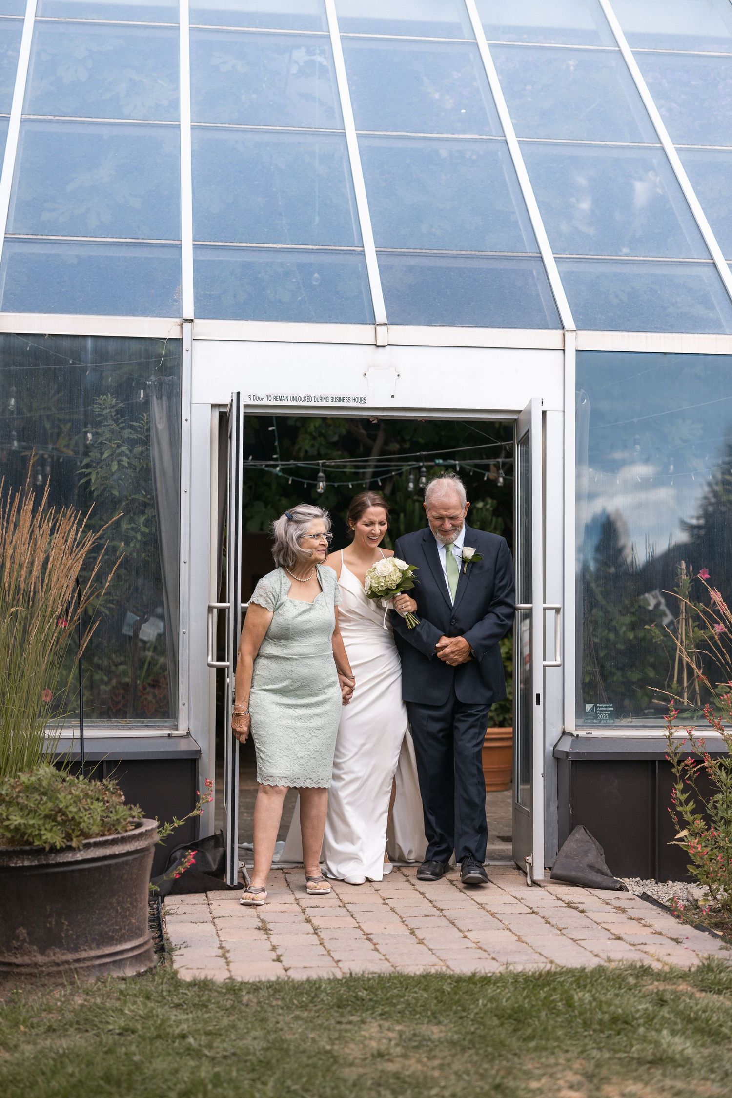 Wedding group emerges from a glass-walled greenhouse during celebration.