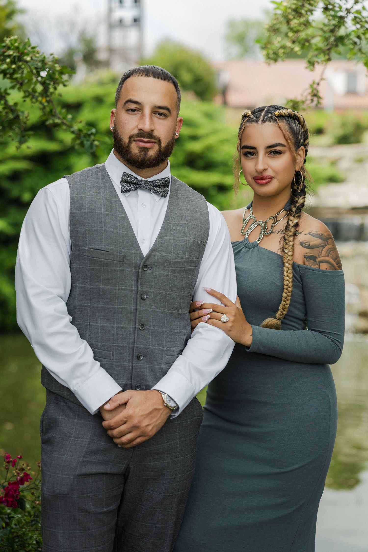 Couple in coordinating gray formal attire posing outdoors near a pond, woman in off-shoulder gown, man in vest and bow tie.