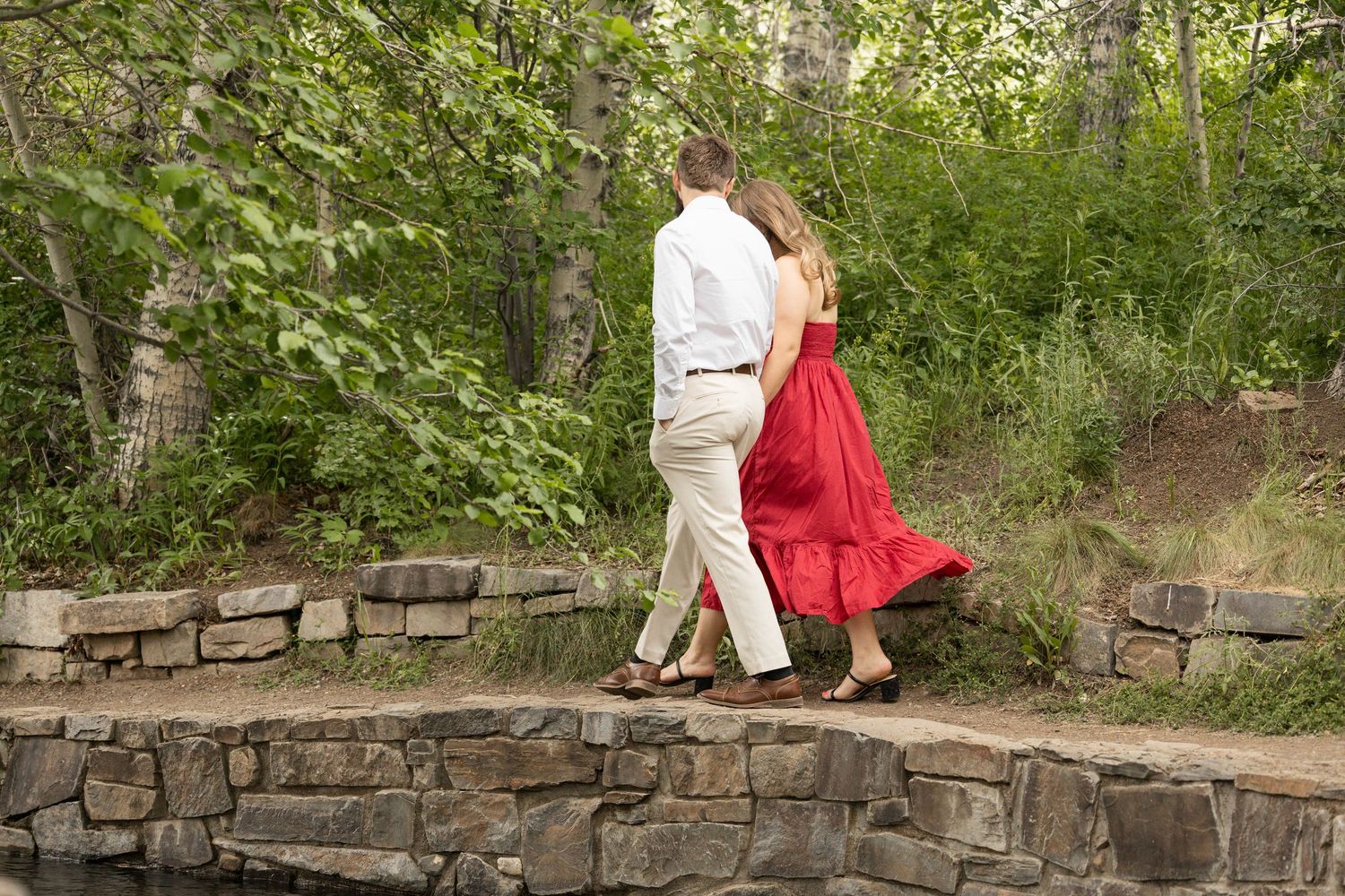 A couple walks romantically along a curved stone wall path surrounded by lush greenery.