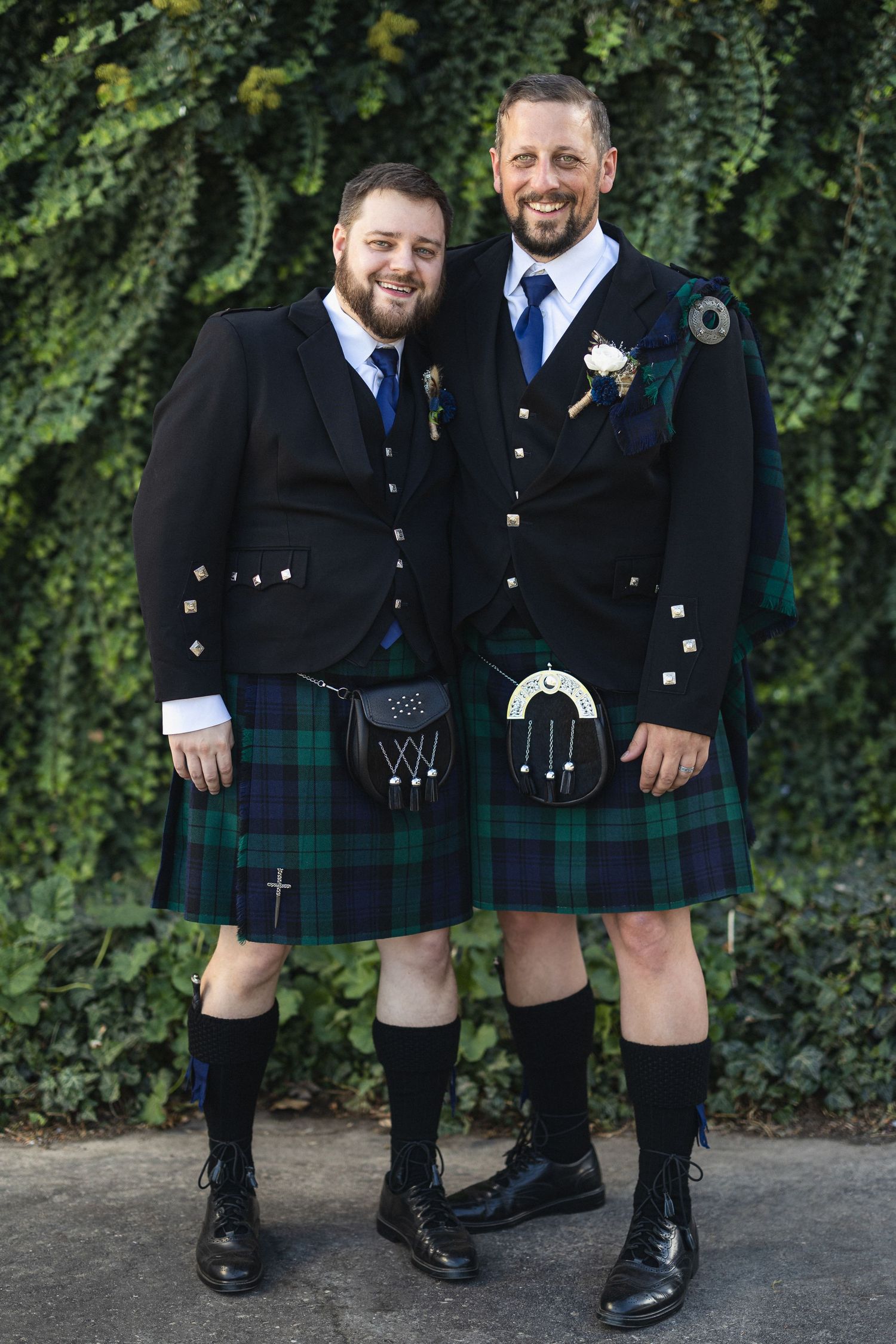 Groomsmen in matching kilts and formal jackets pose against green ivy background.