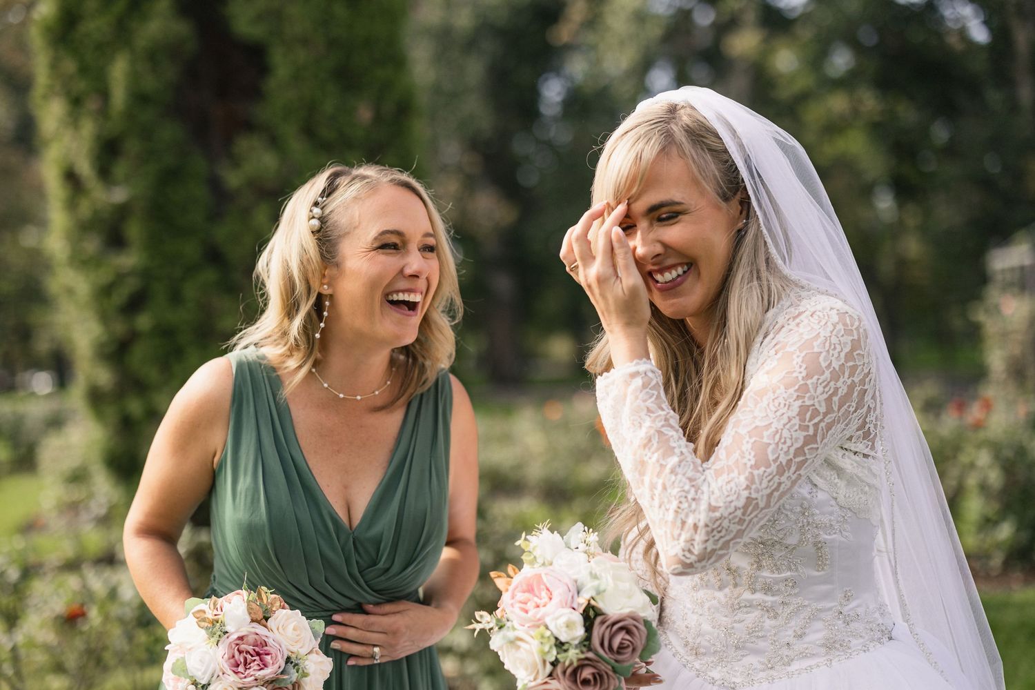 Two women share genuine laughter while holding pink and white bouquets during wedding celebrations.