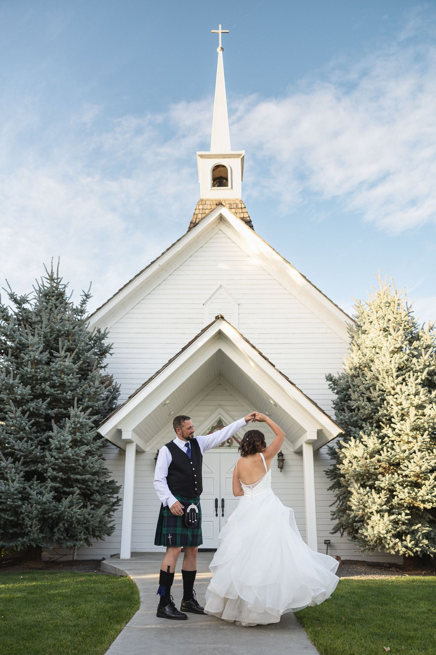 A couple poses in front of a charming white country church with a tall steeple on their wedding day.