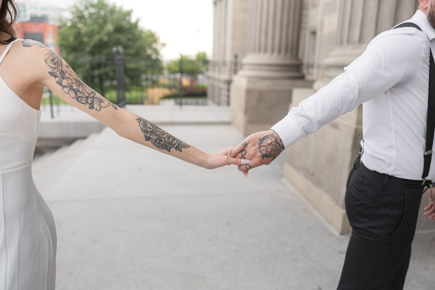 Close-up of tattooed arms as a couple holds hands in front of a classic stone building.