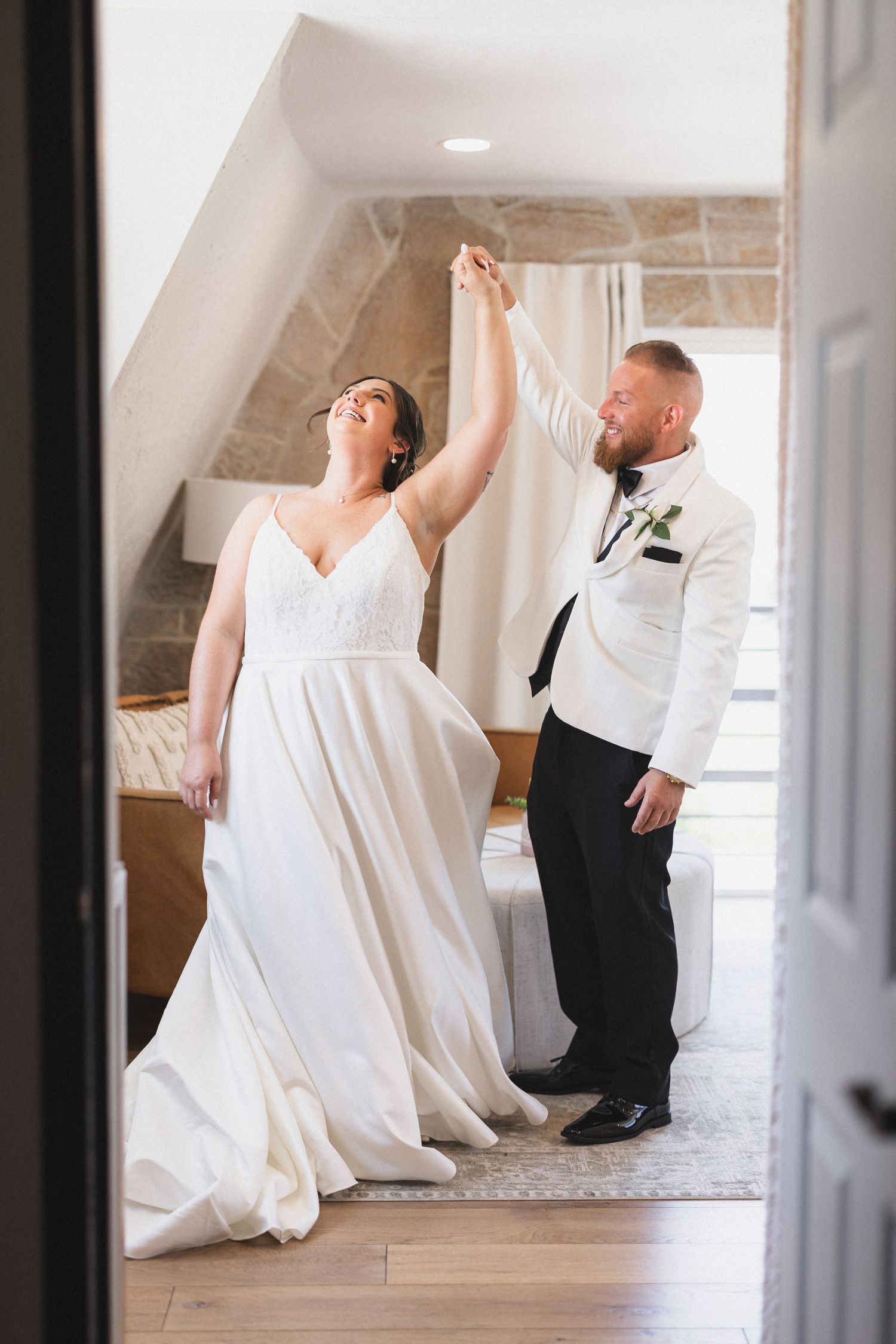 A couple dancing together in a bright hallway, with the bride wearing a flowing white gown and groom in a white tuxedo jacket.