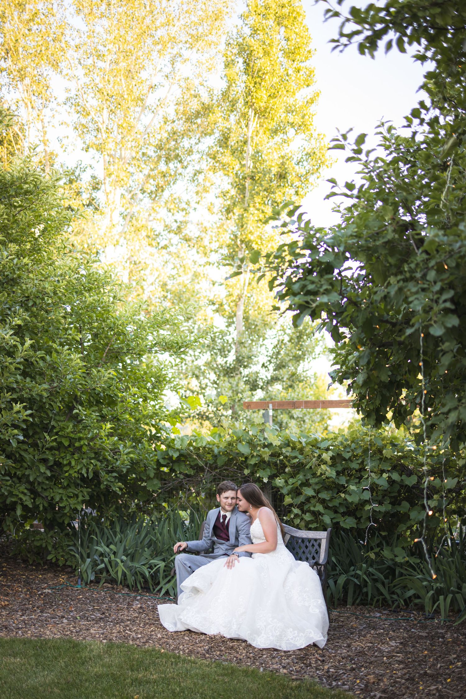 A couple in wedding attire sits together on a garden bench surrounded by lush greenery and golden trees.