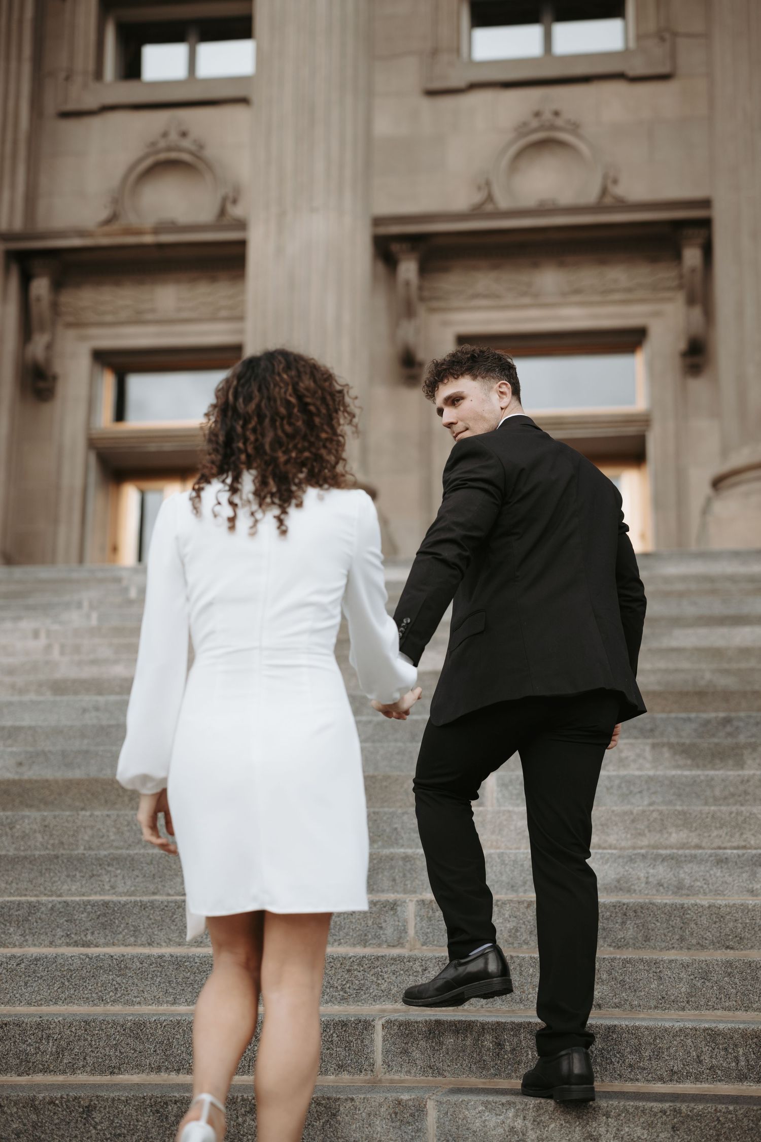 A couple climbs stone steps together while holding hands in formal attire.