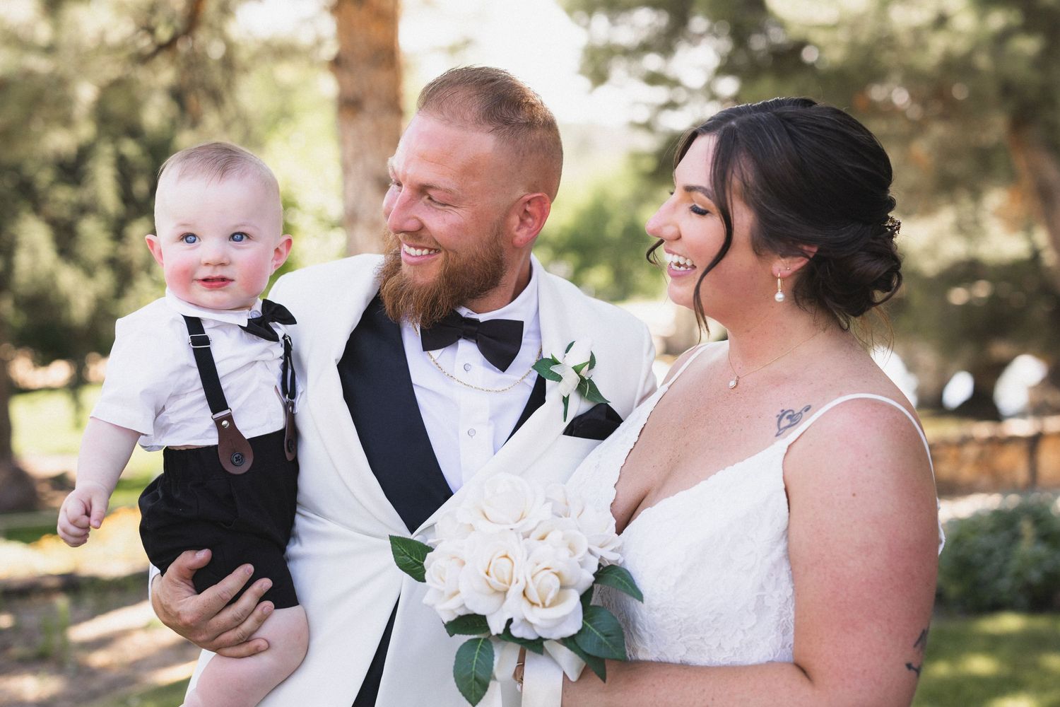 Joyful wedding portrait with couple and baby in formal attire against outdoor backdrop.