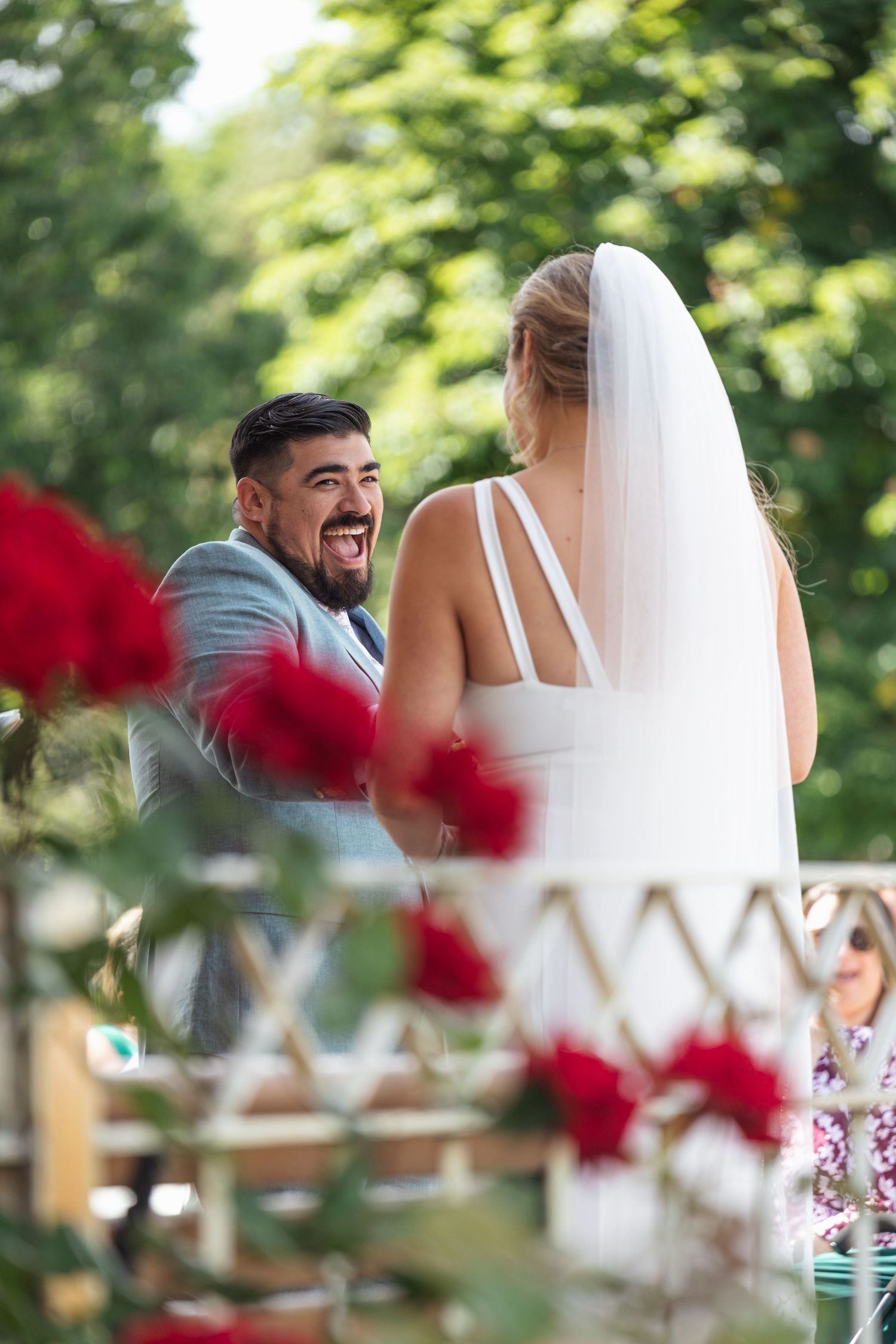 Couple shares a joyful moment during outdoor wedding ceremony with red roses in foreground.