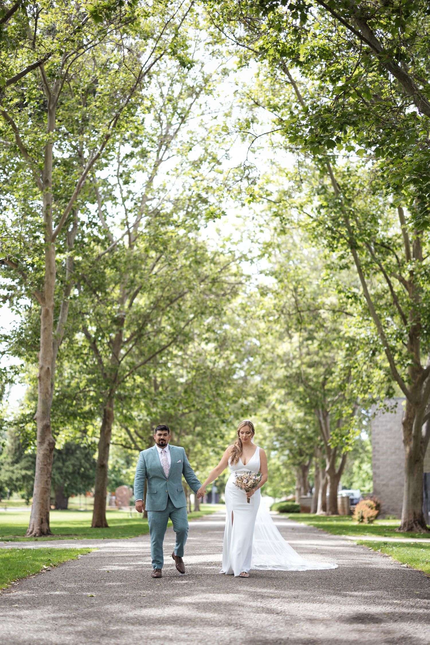 Newlyweds walk down tree-lined path during outdoor wedding ceremony.