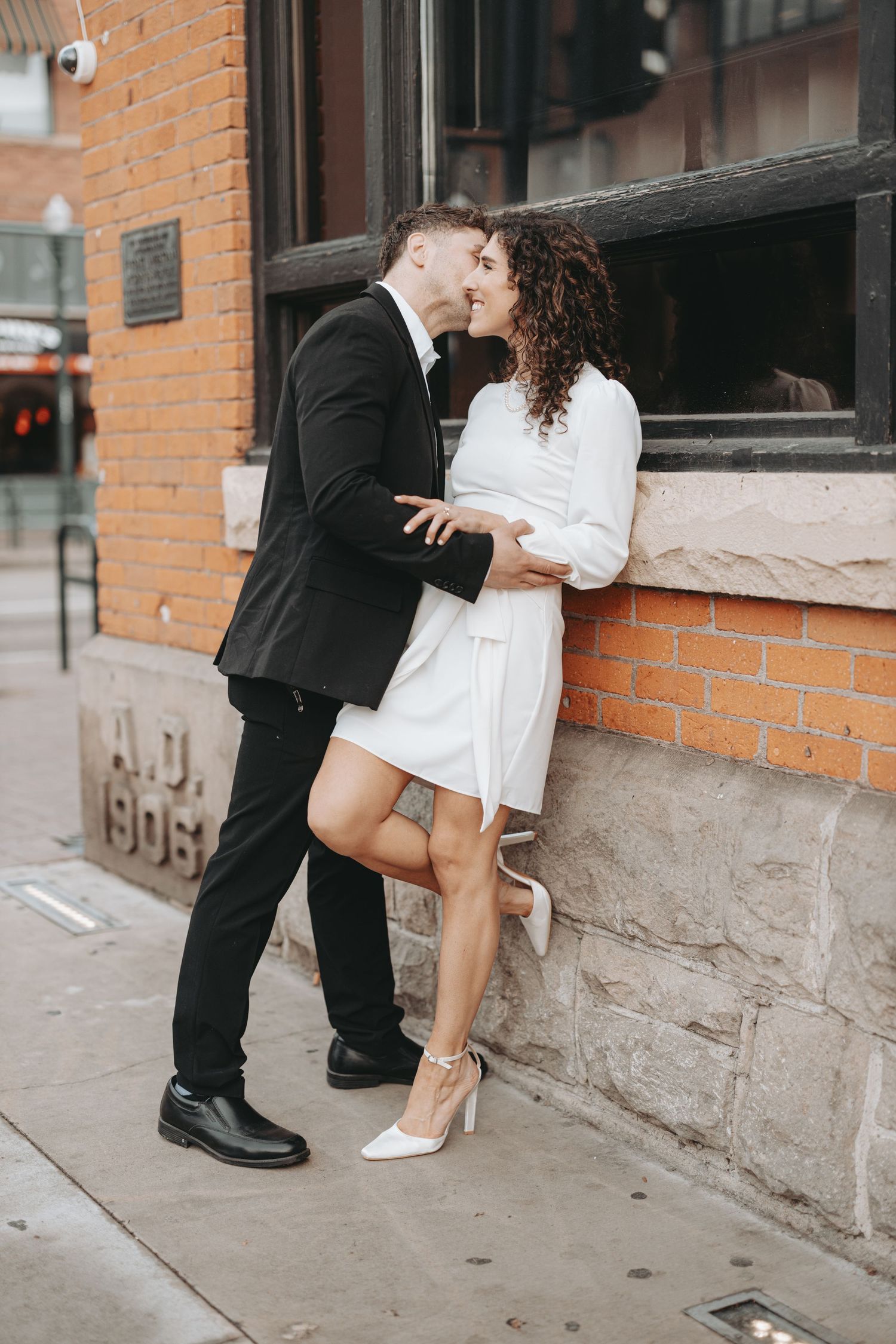 A romantic couple sharing an intimate moment against a brick wall, she in white dress and he in black suit.