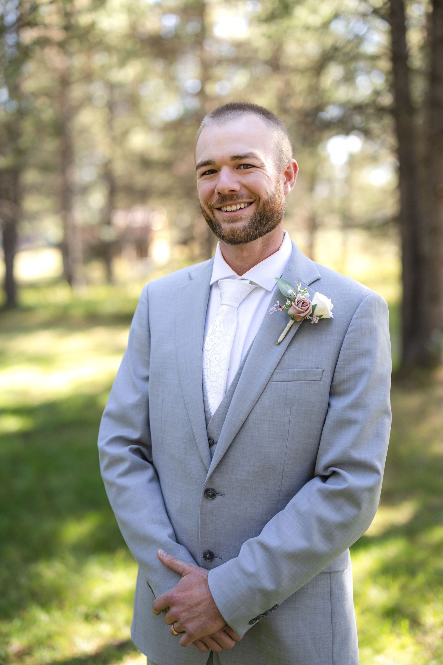 Groom in light gray suit with boutonniere smiling in an outdoor forest setting.