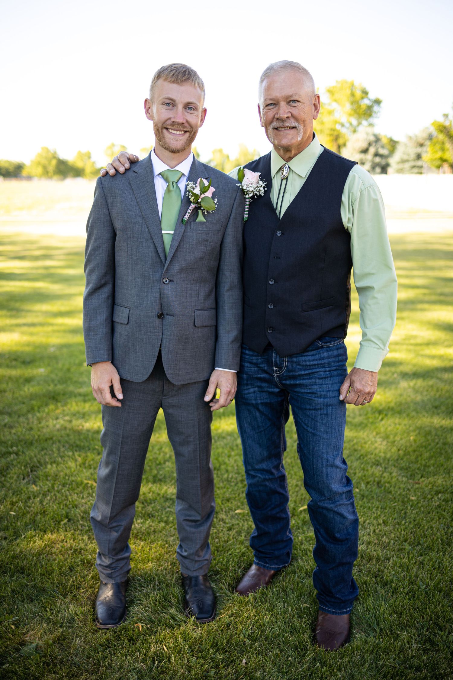 Two men in formal wear with matching green accents pose on sunny lawn.