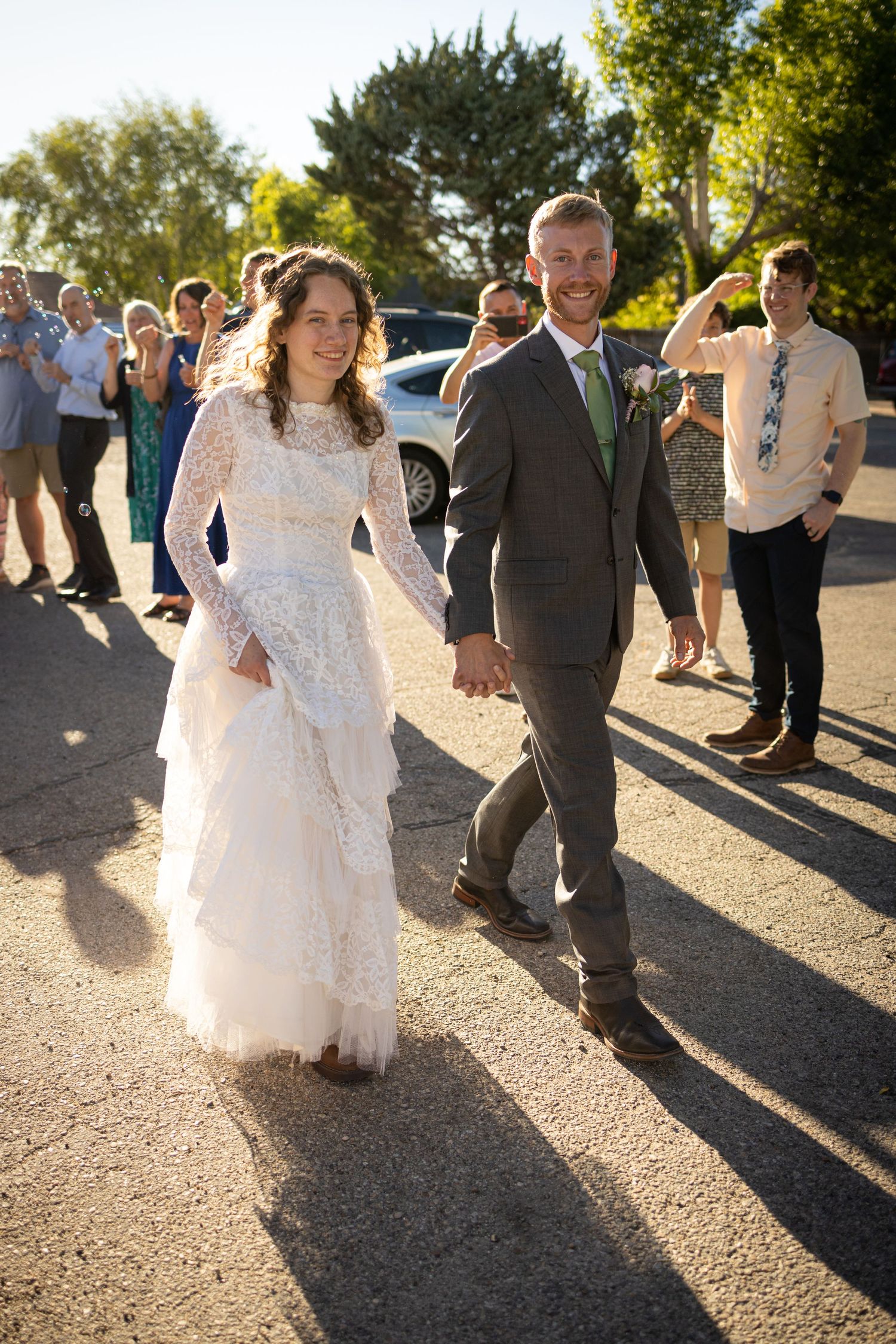 Newlyweds walking together outdoors with wedding guests cheering in the background.