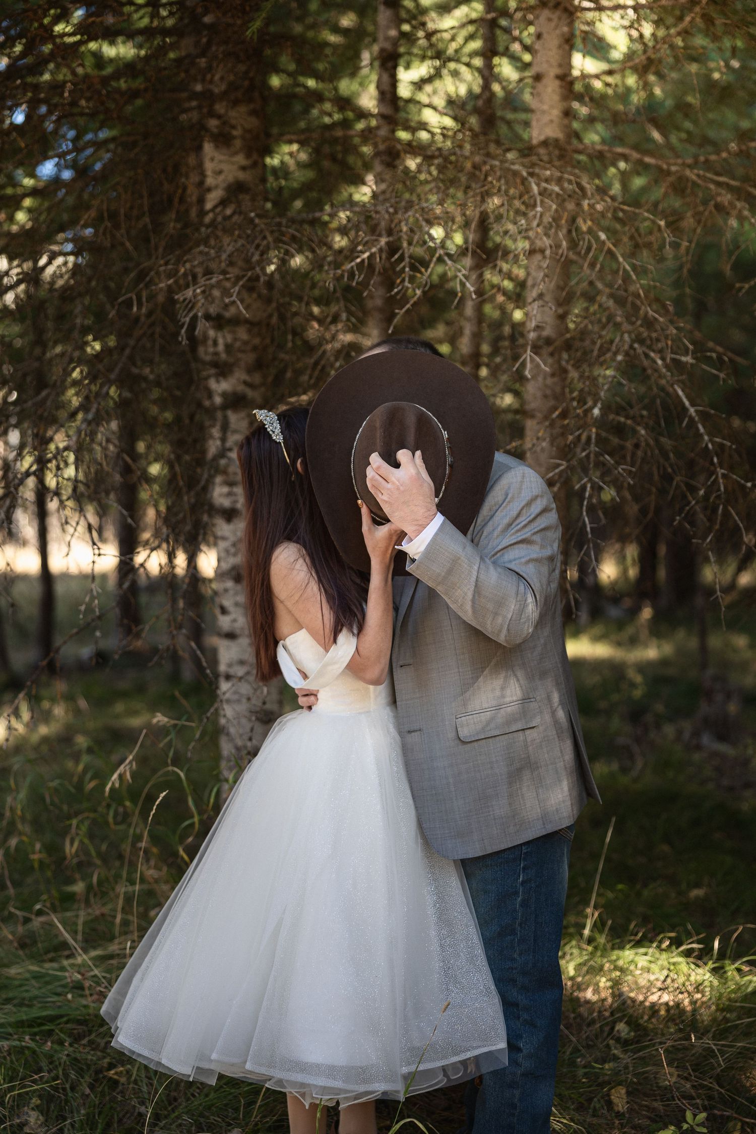 A couple in wedding attire share a private laugh while standing in a wooded area.