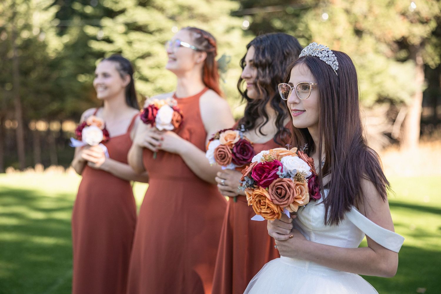 Bridal party in terracotta dresses holds floral bouquets while standing on green lawn.