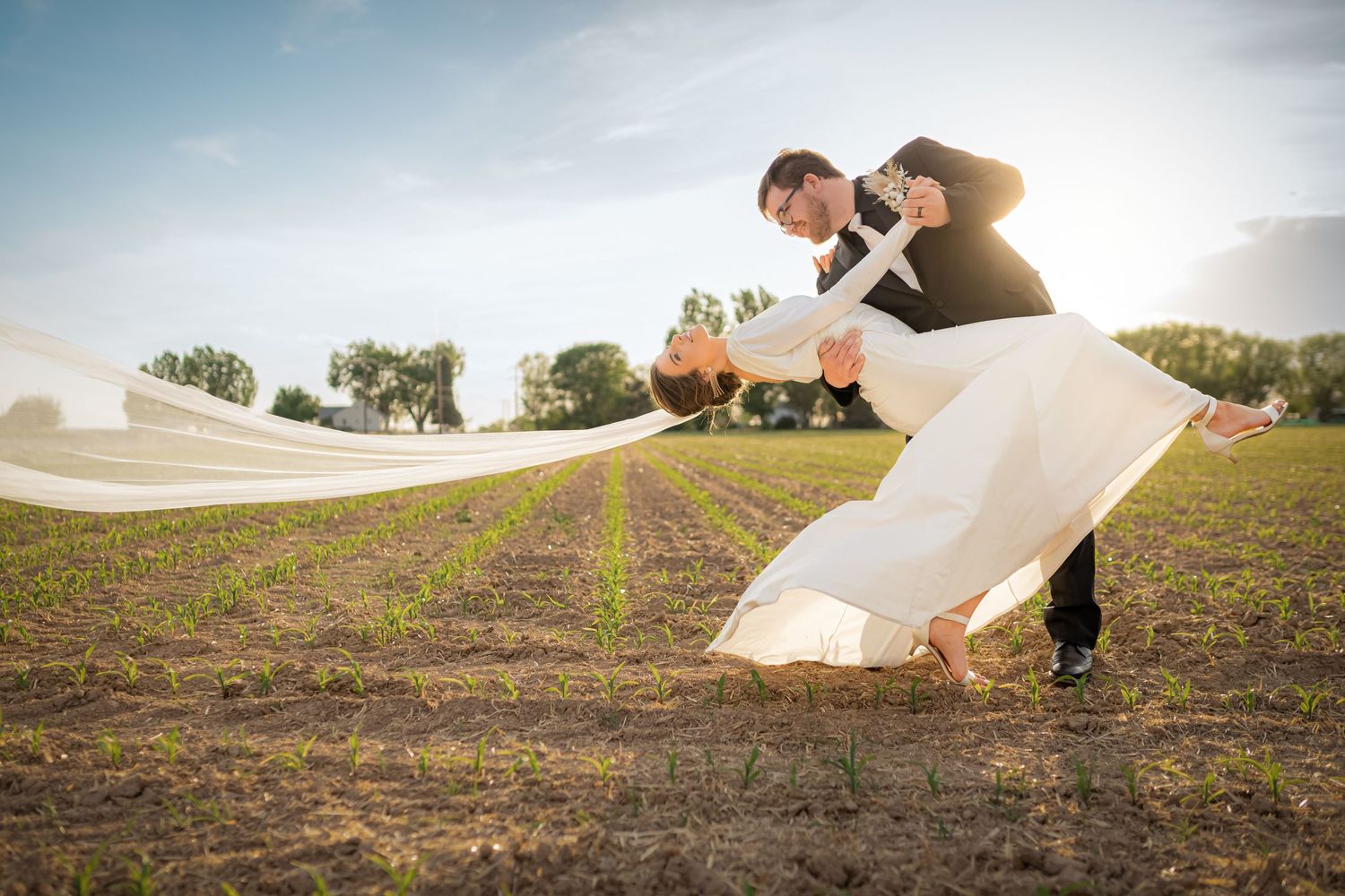 Dramatic bridal couple portrait with flowing veil in rustic outdoor setting at sunset.