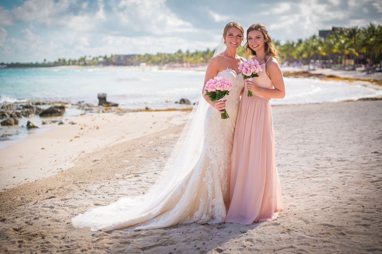 Two people in formal attire pose together on a tropical beach with palm trees and turquoise water in the background.