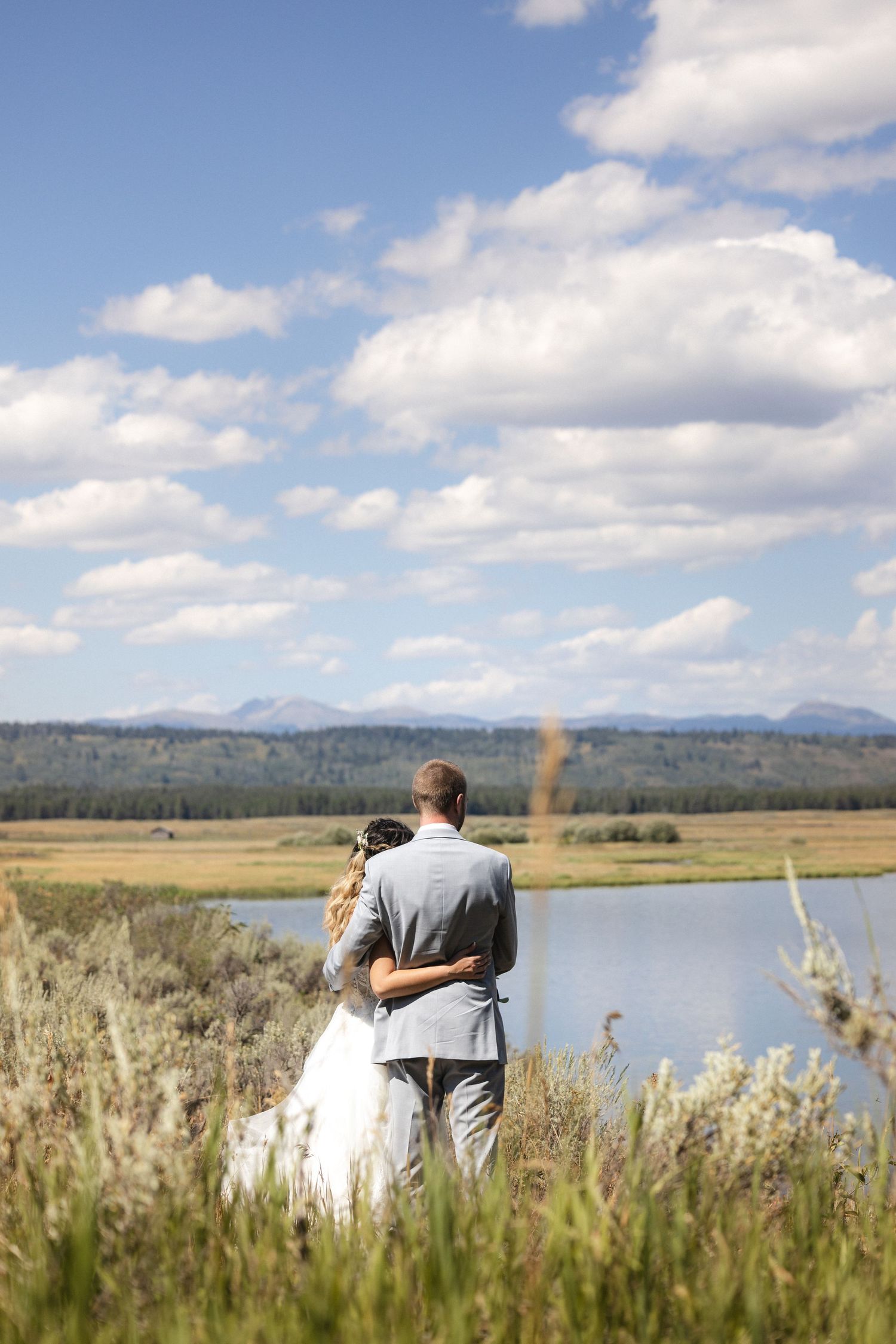 Wedding couple stands near a serene lake with mountains and blue sky in the background.