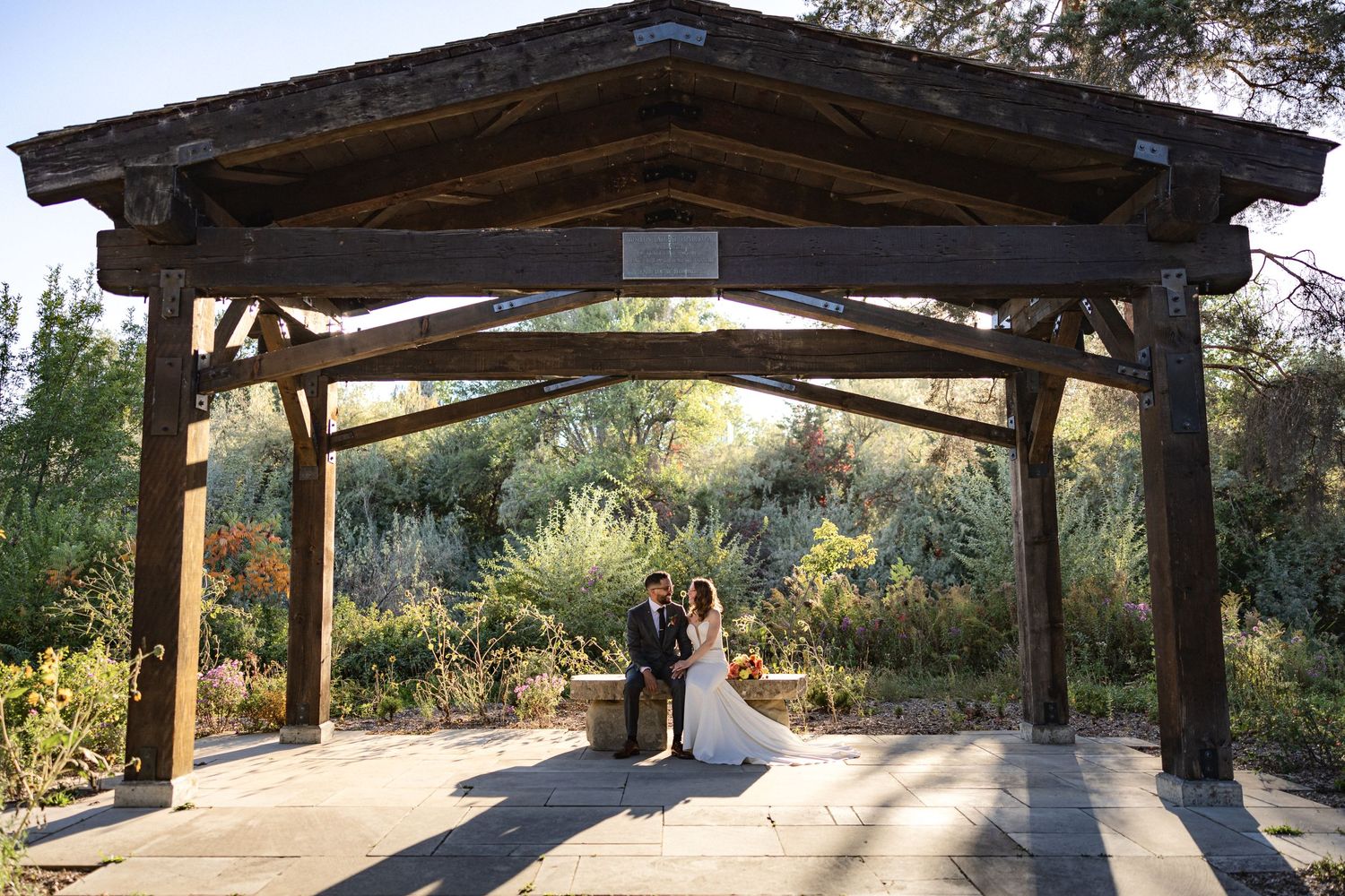 Bride and groom stand under wooden pavilion surrounded by lush garden landscaping.