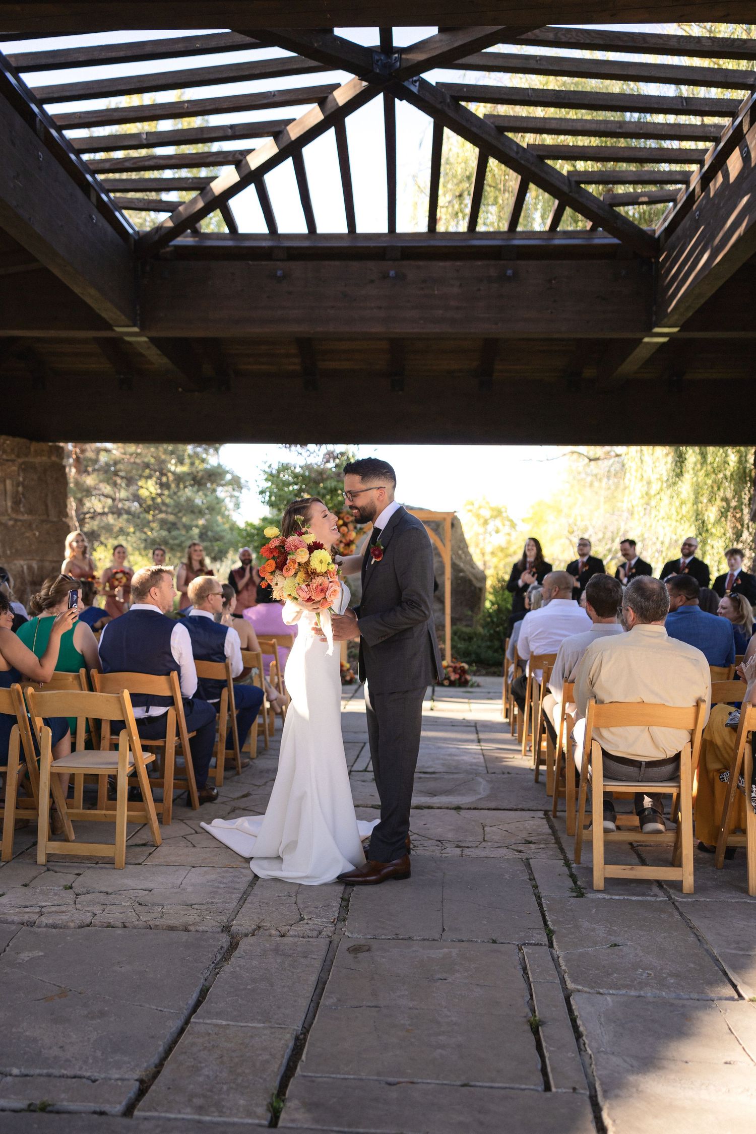 Outdoor wedding ceremony under a wooden pergola with guests seated on wooden folding chairs.