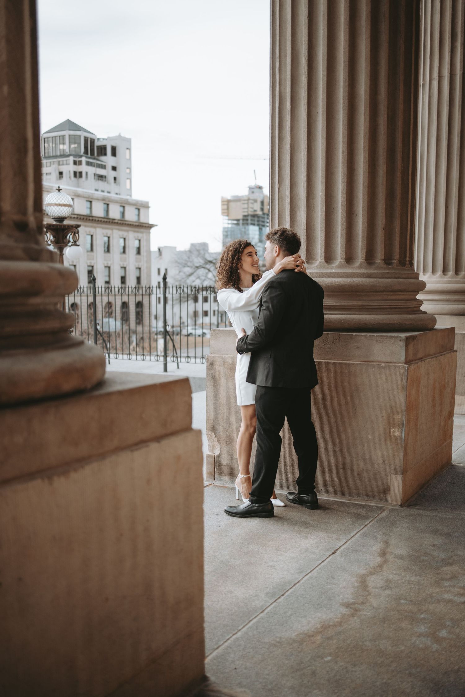A couple embraces between grand stone columns with city architecture visible in background.