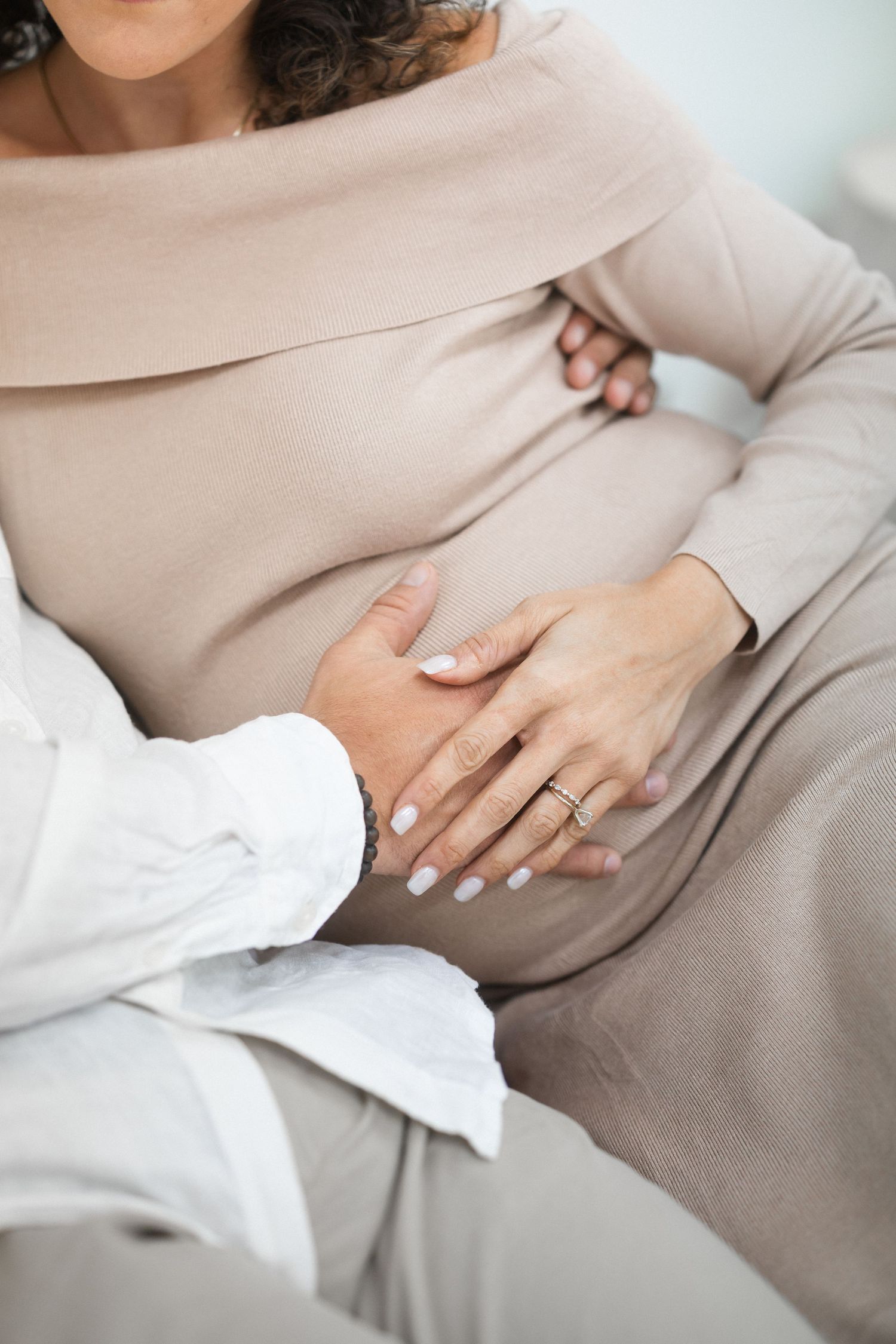 Close-up view of hands resting on a pregnant belly wearing a beige sweater and white sleeves.