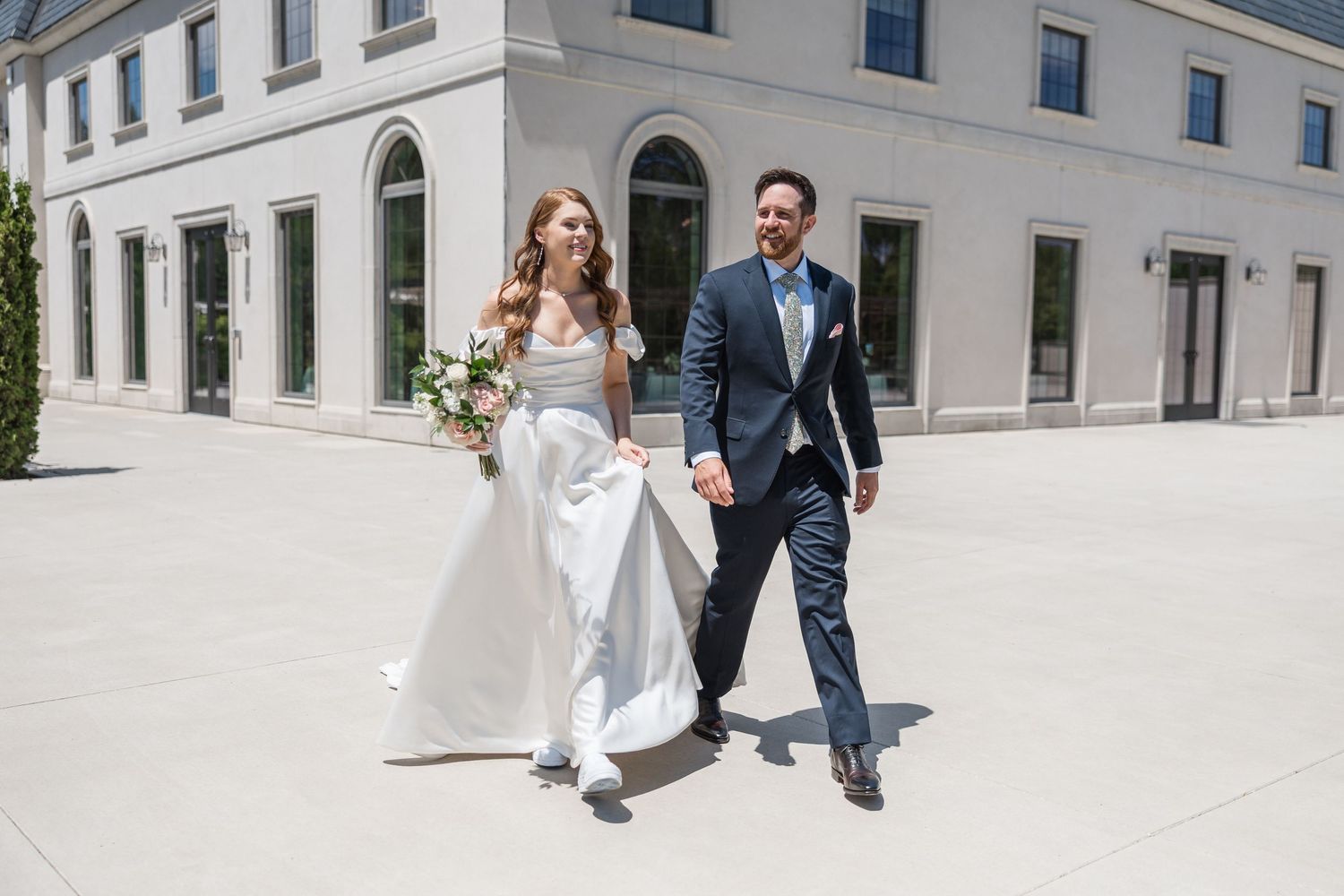 Newlyweds walking together outside elegant white building on sunny day.