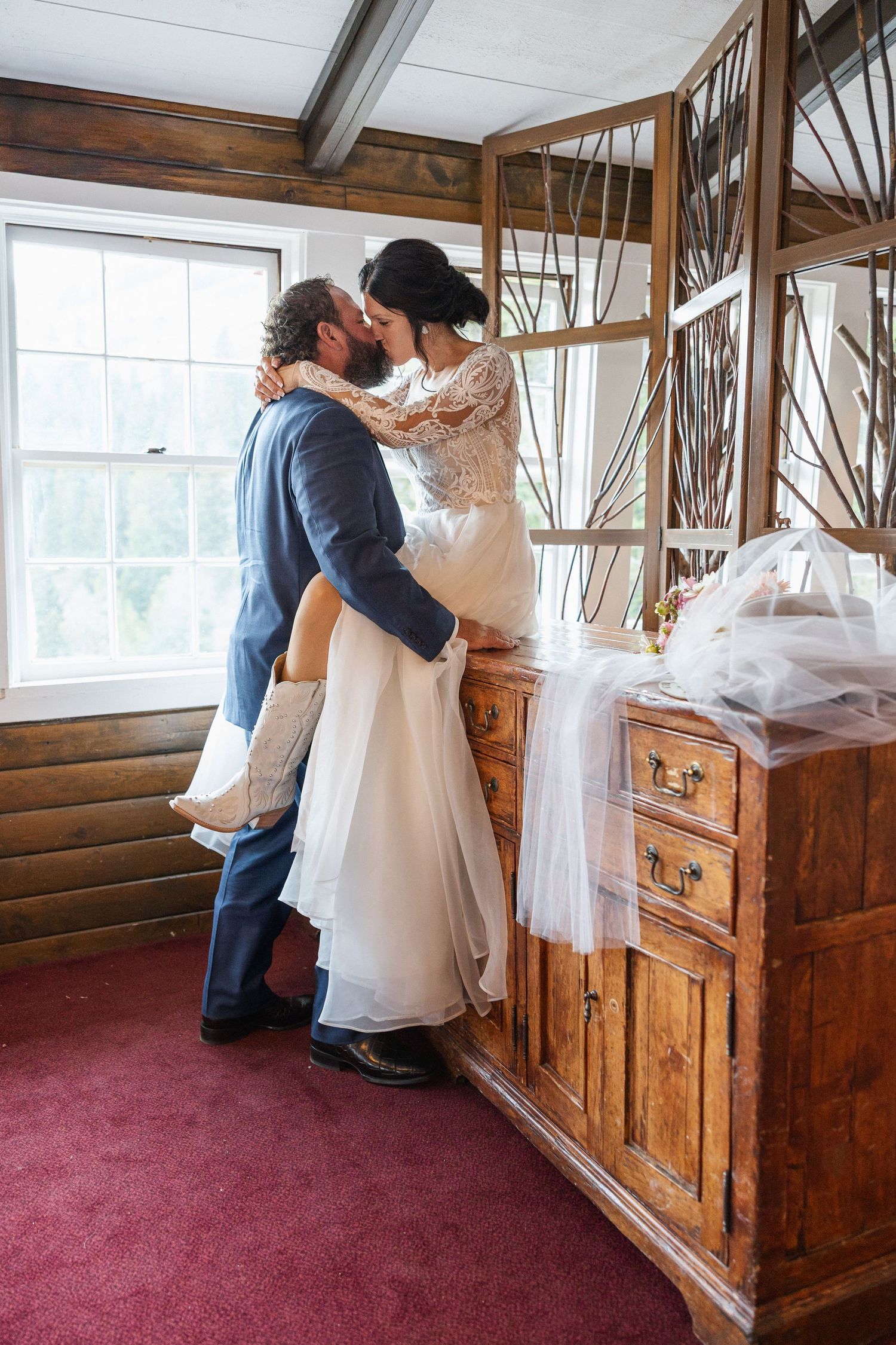 A couple embraces in front of a rustic wooden dresser with a mirror during a wedding celebration.