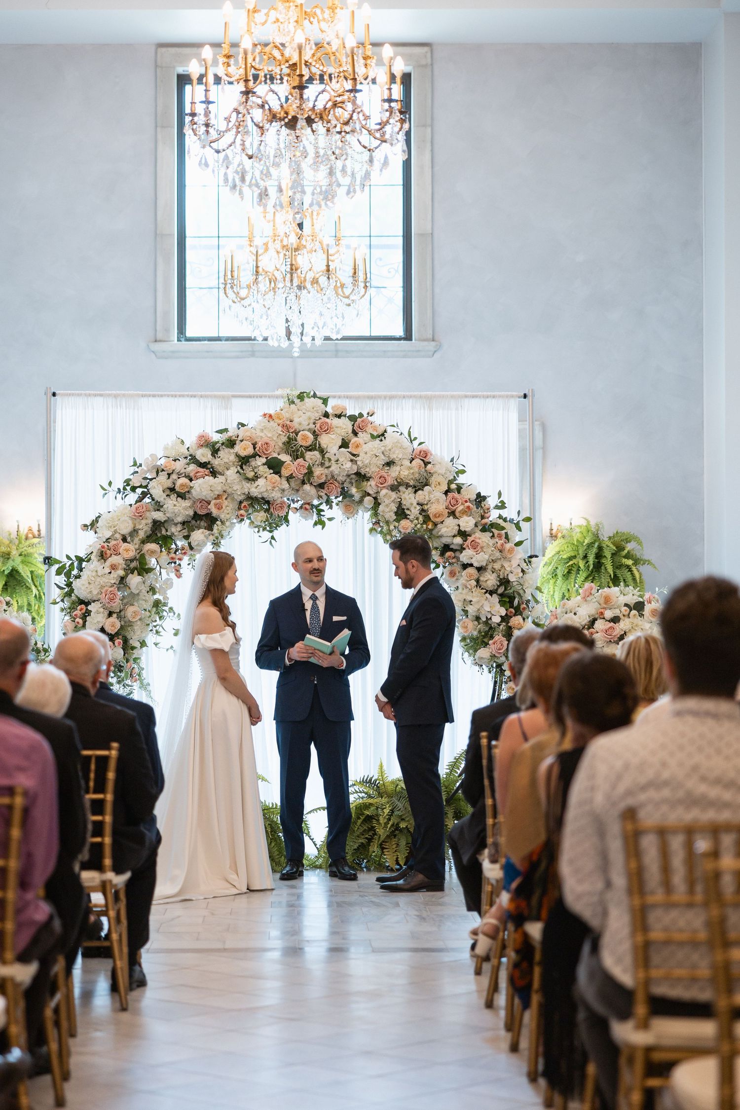 Indoor wedding ceremony under crystal chandelier with floral arch and seated guests.