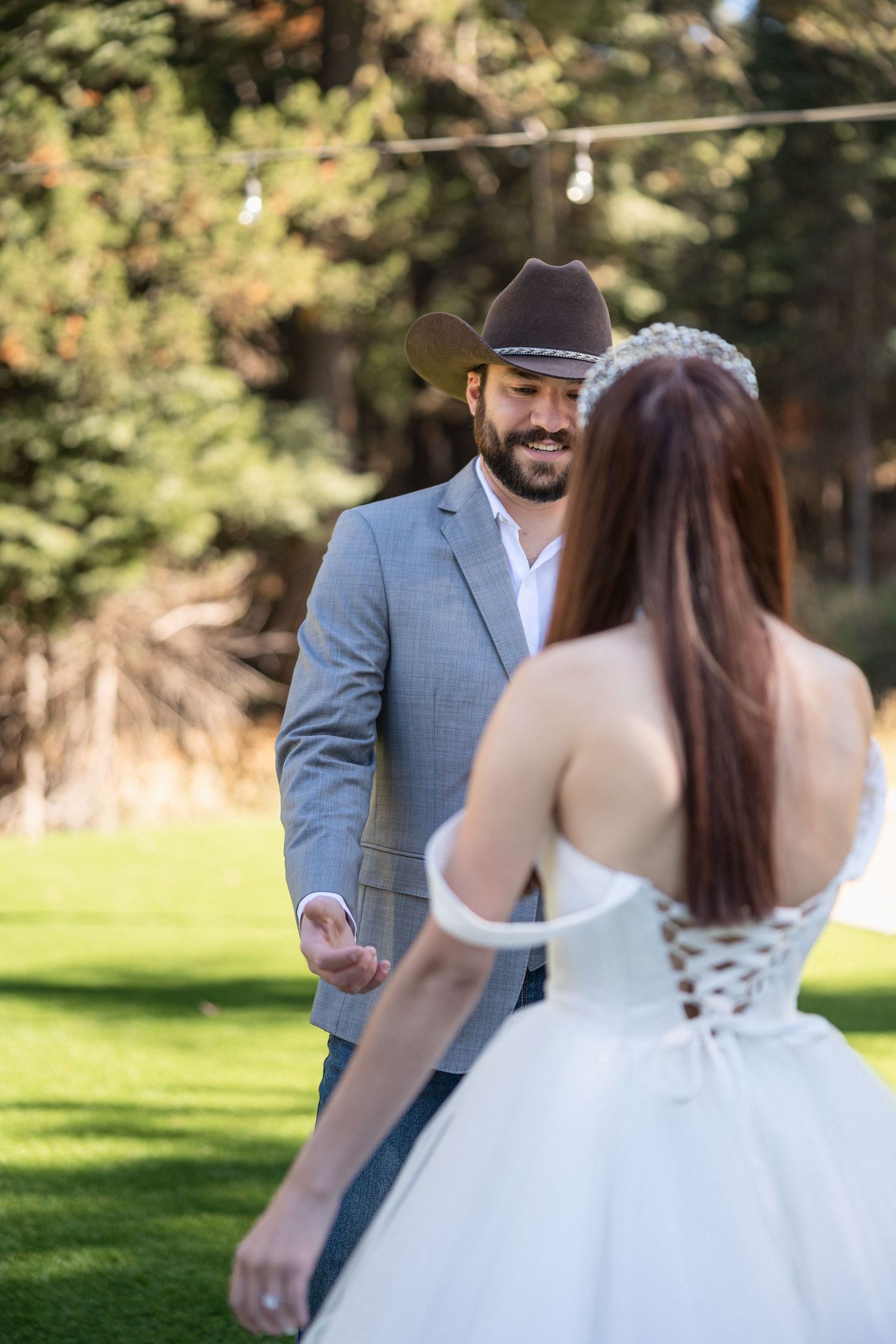 A couple dances together outdoors, with the bride wearing a white ballgown and the groom in a grey suit and cowboy hat.