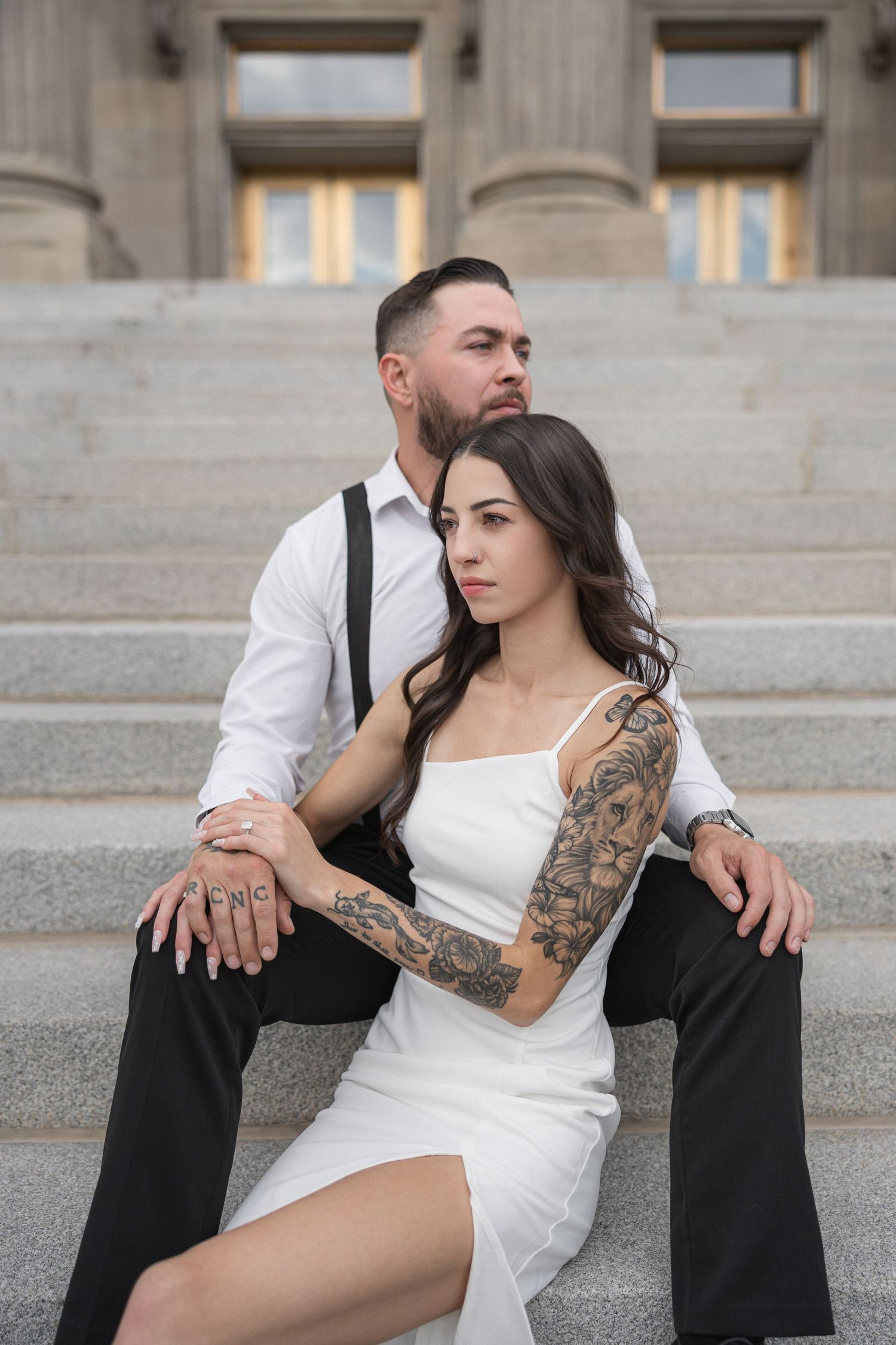 Couple in formal attire poses on stone steps with visible tattoos.