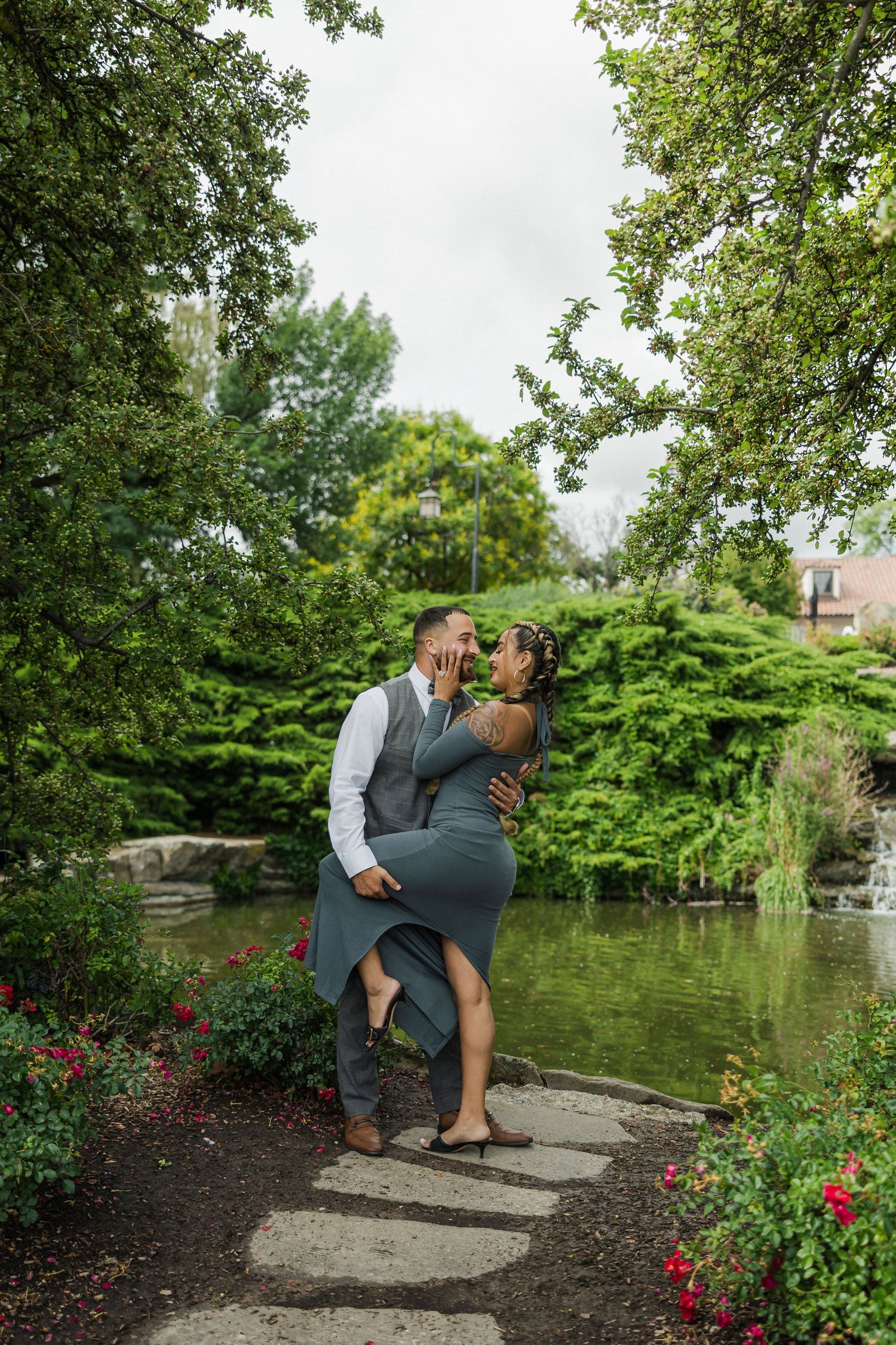 A couple shares a romantic dip kiss on stepping stones beside a pond in a lush green garden.