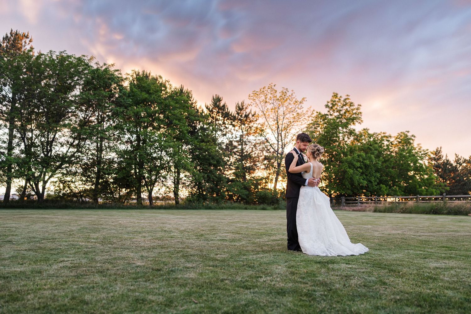 Bride and groom share a romantic kiss at sunset on a lush green lawn with colorful pink and orange skies.