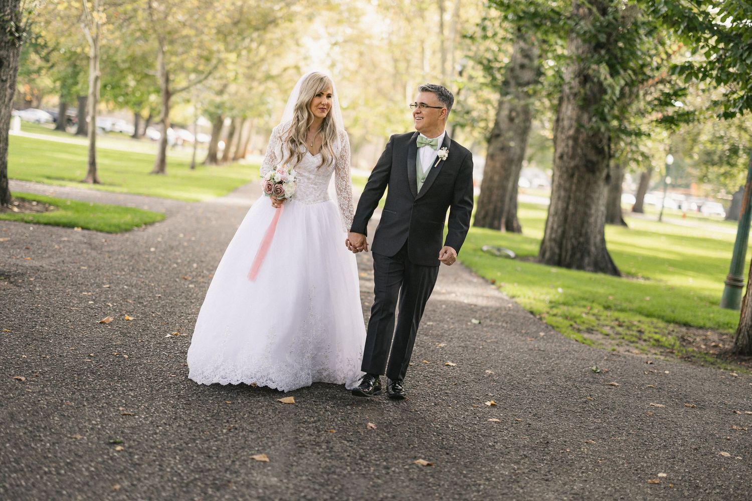 Couple walking hand in hand along a tree-lined path in formal wedding attire.
