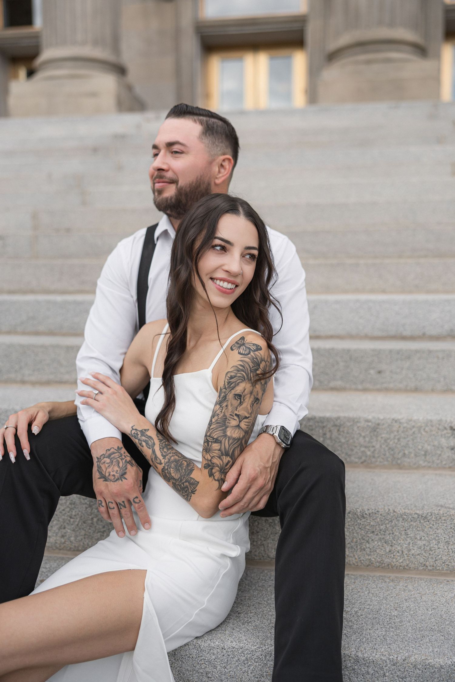 A couple in formal wear sitting on stone steps, showcasing intricate tattoo artwork on their arms.